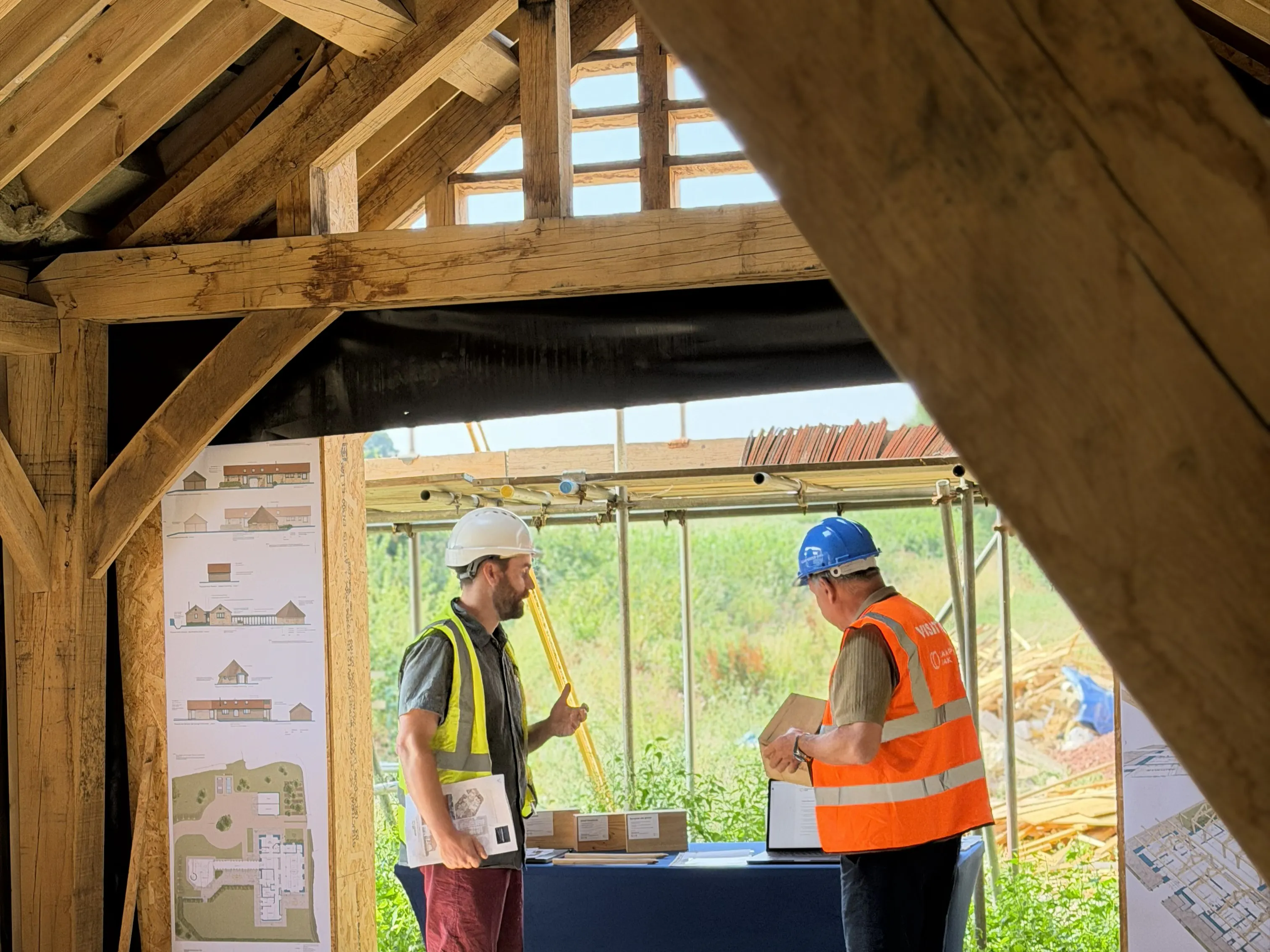 An event on a construction site where attendees in high vis jackets and blue hard hats are shown around an oak frame house mid-build