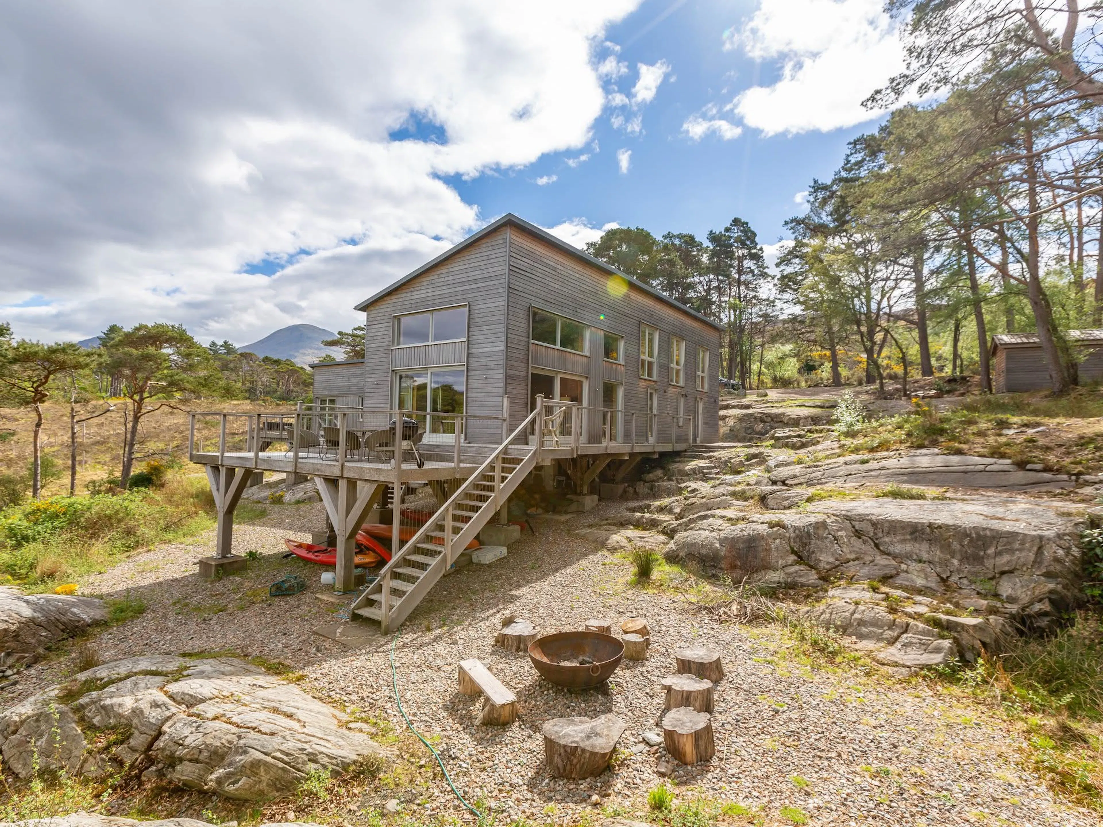 Exterior view of a timber clad Douglas fir framed house beside a loch with a rocky shoreline, and a fire pit on the beach in the foreground