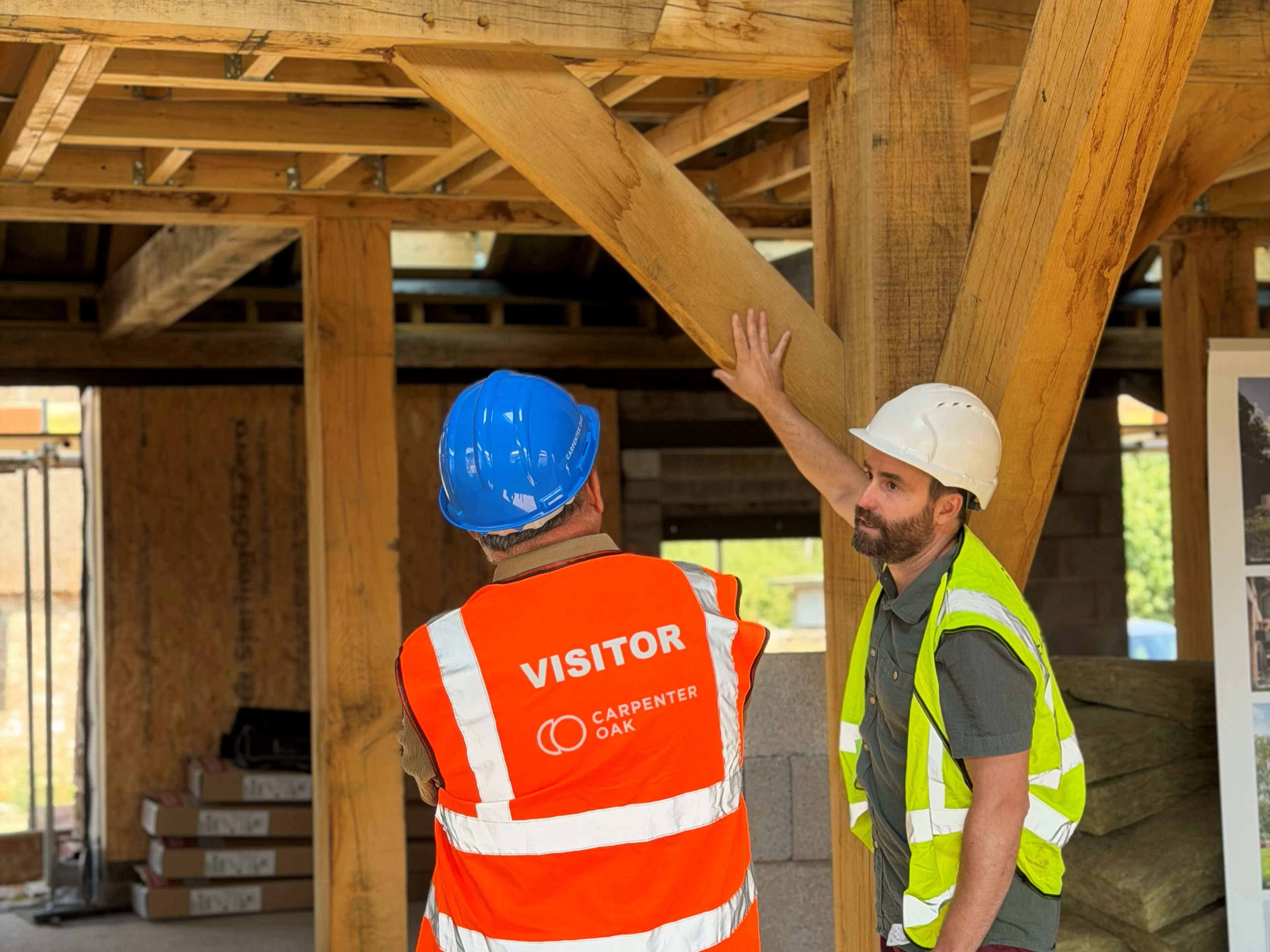 An event on a construction site where attendees in high vis jackets and blue hard hats are shown around an oak frame house mid-build