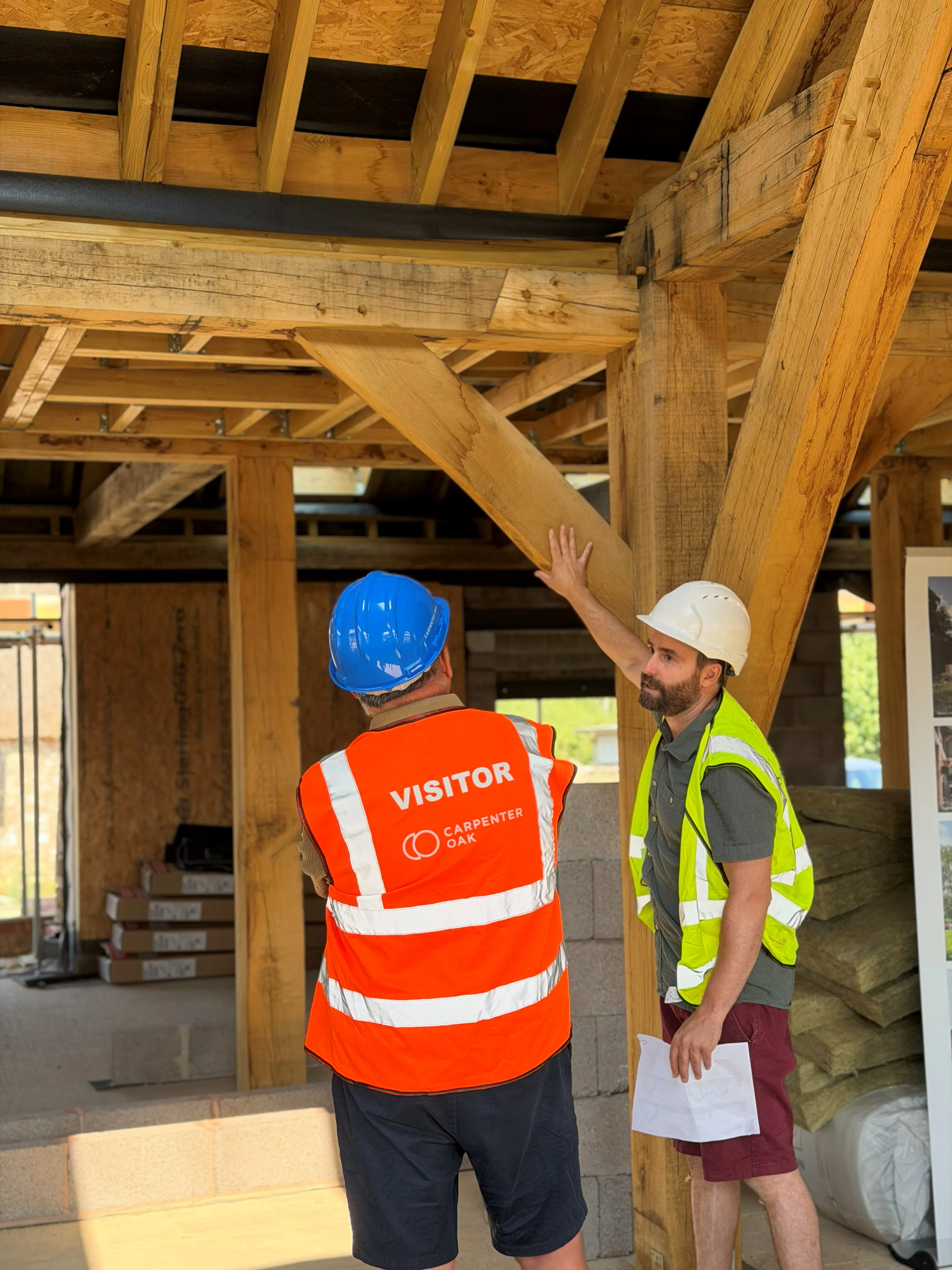 An event on a construction site where attendees in high vis jackets and blue hard hats are shown around an oak frame house mid-build