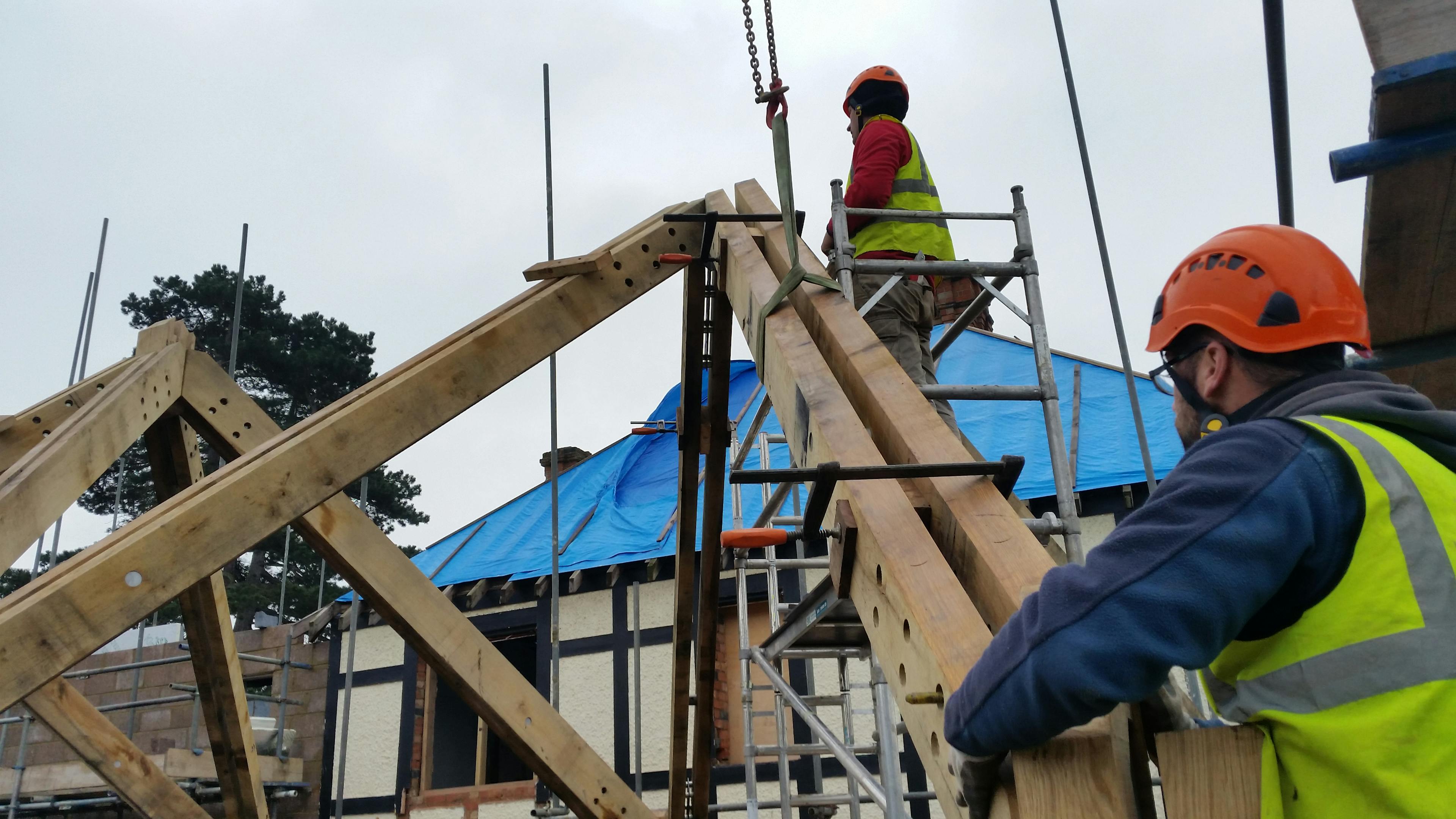 Two carpenters with hard hats and high vis jackets install an oak frame with steel connections