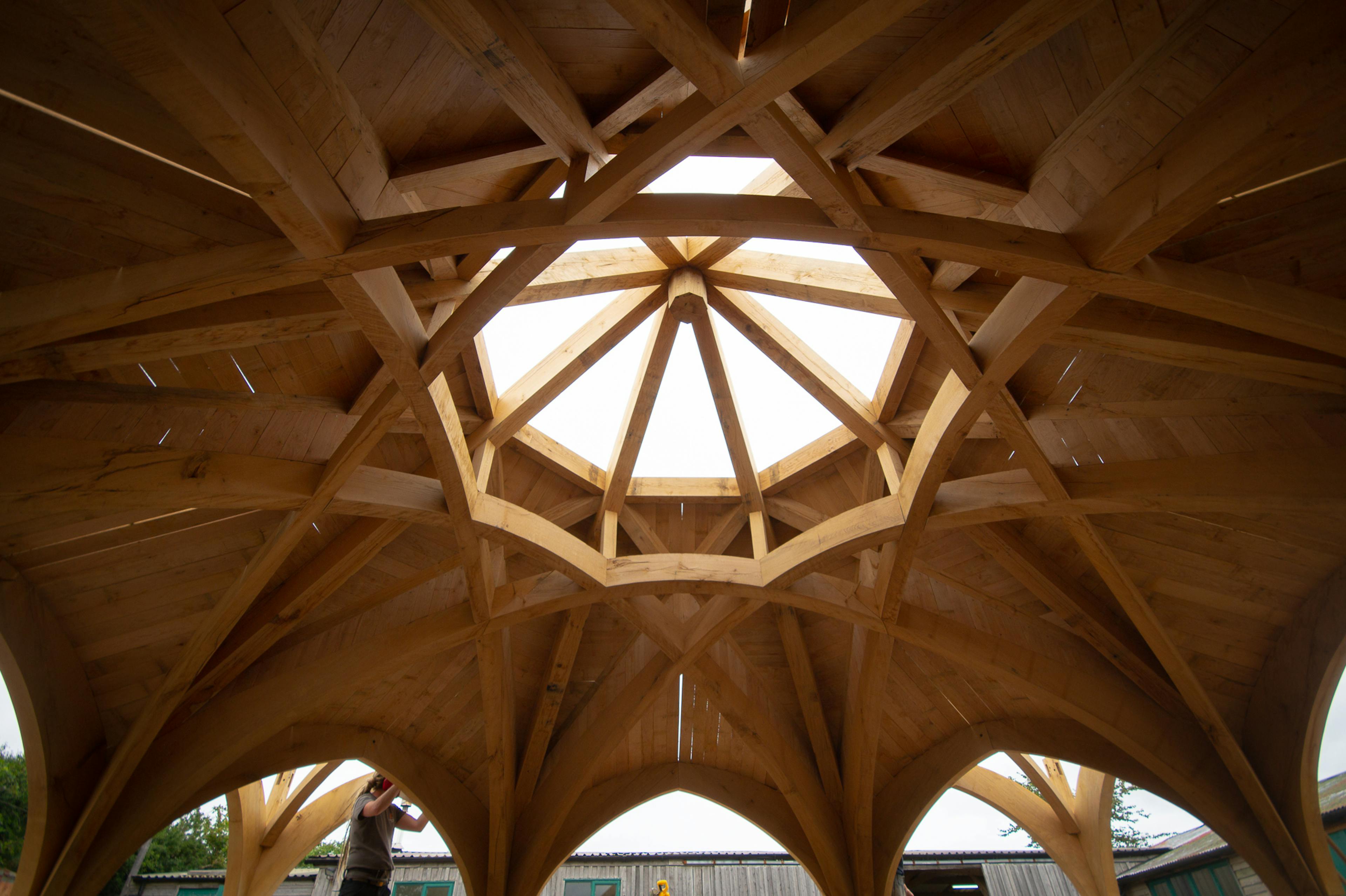The underneath of a complex octagonal oak roof structure outside some workshops in a carpentry yard