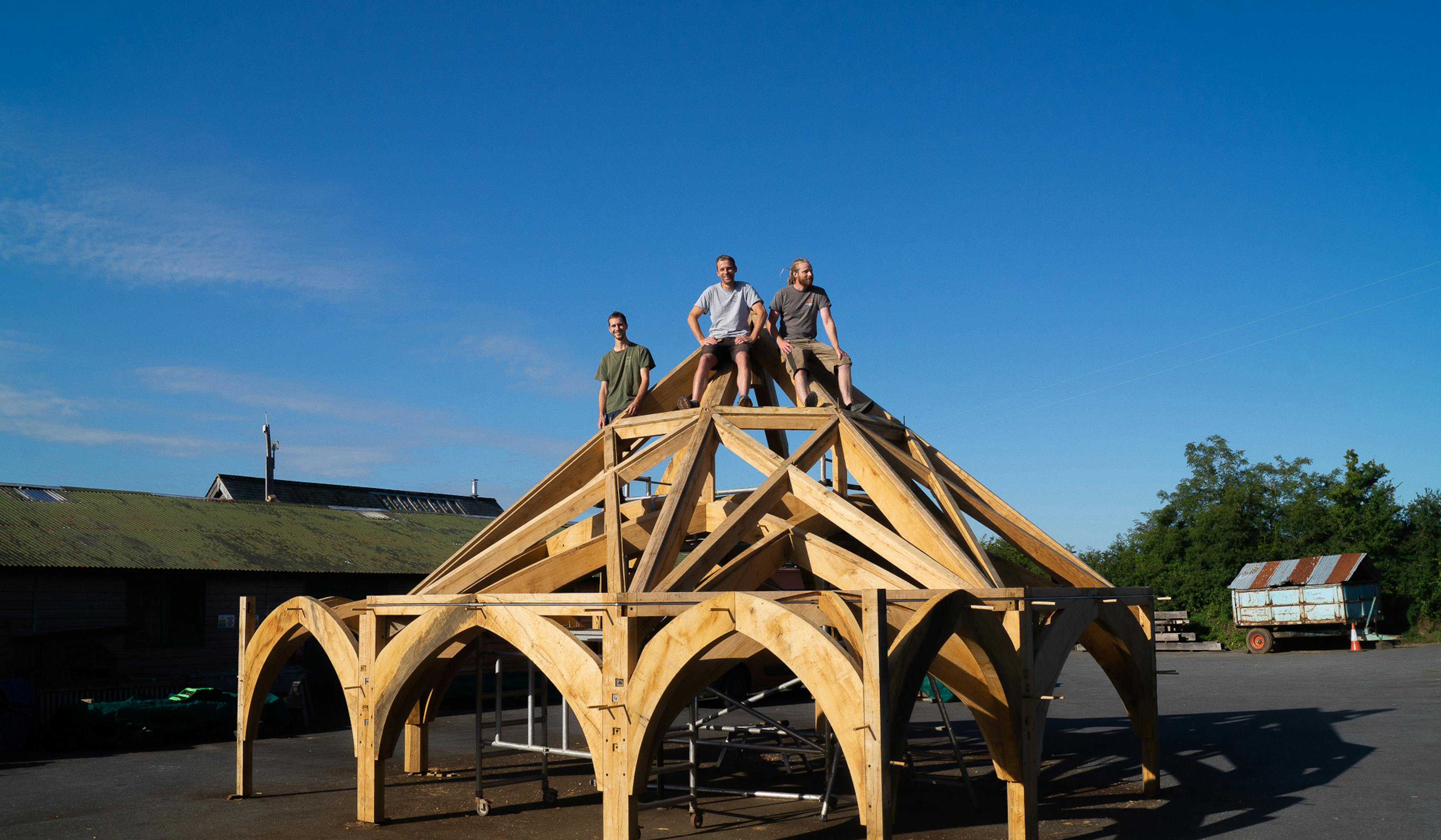 Three carpenters sit on a complex octagonal oak roof structure outside some workshops in a carpentry yard