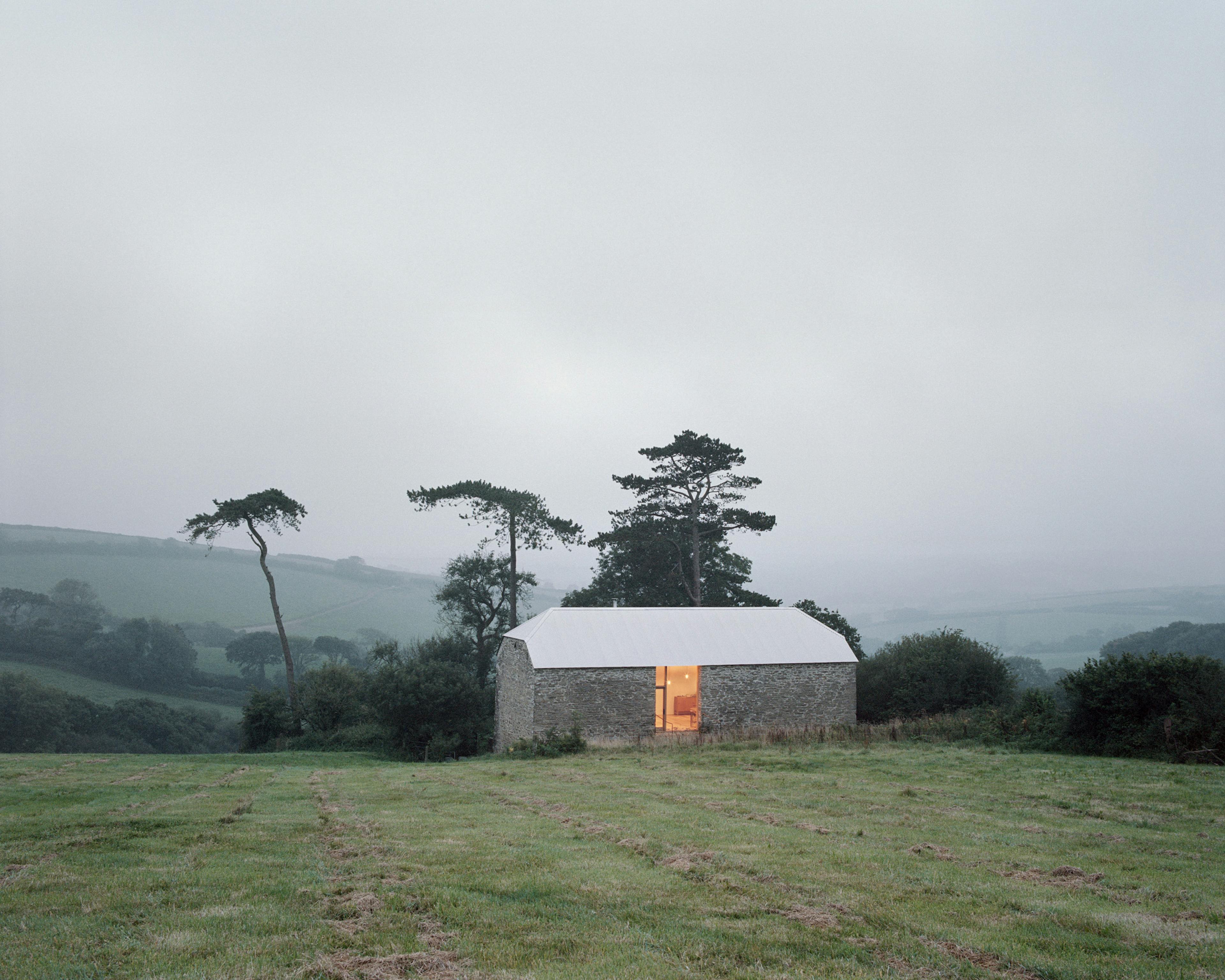 A light shines from the door of a renovated stone barn at dusk in a field