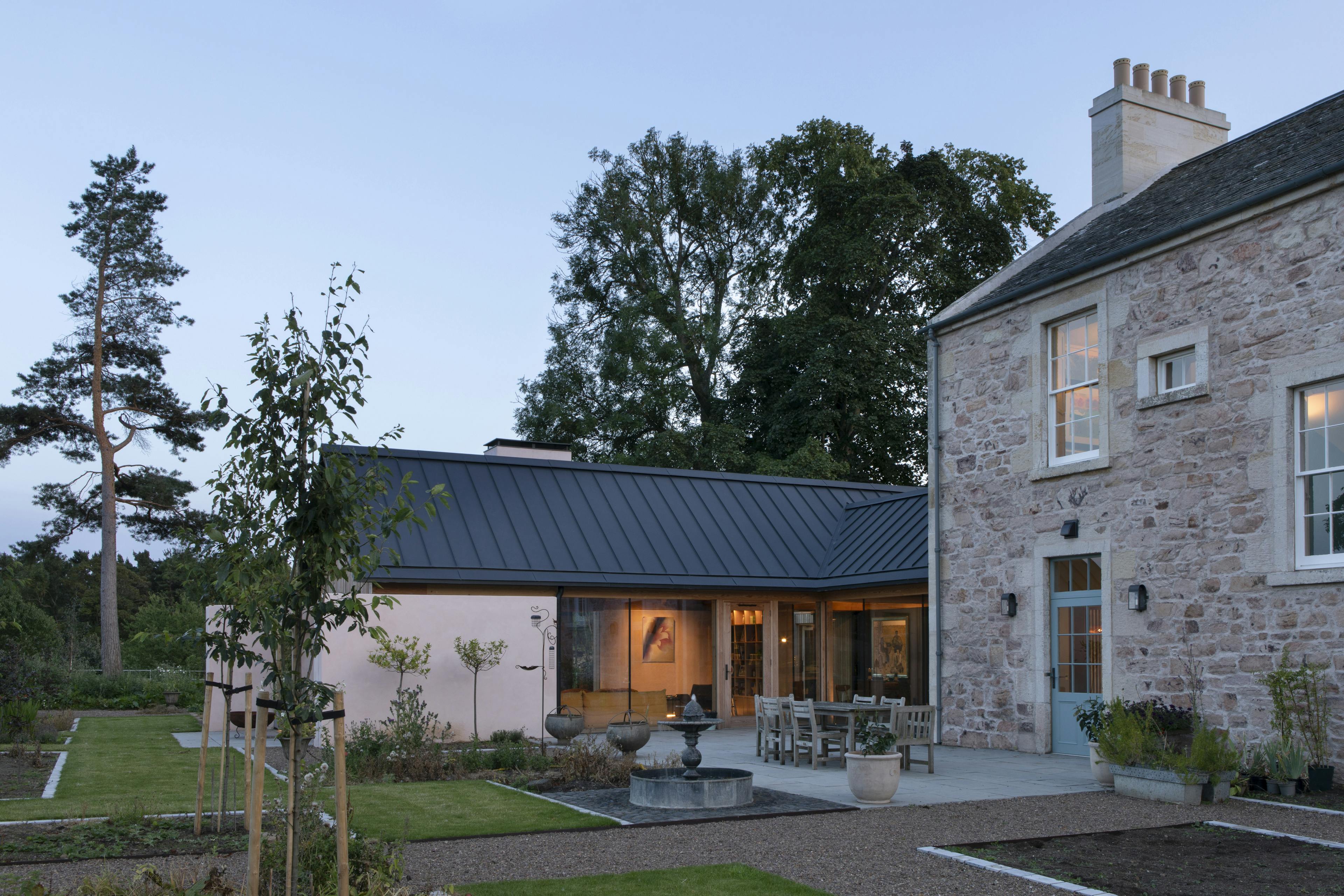 At dusk light shines from a Douglas fir framed extension to a Georgian country manor house set in landscaped gardens