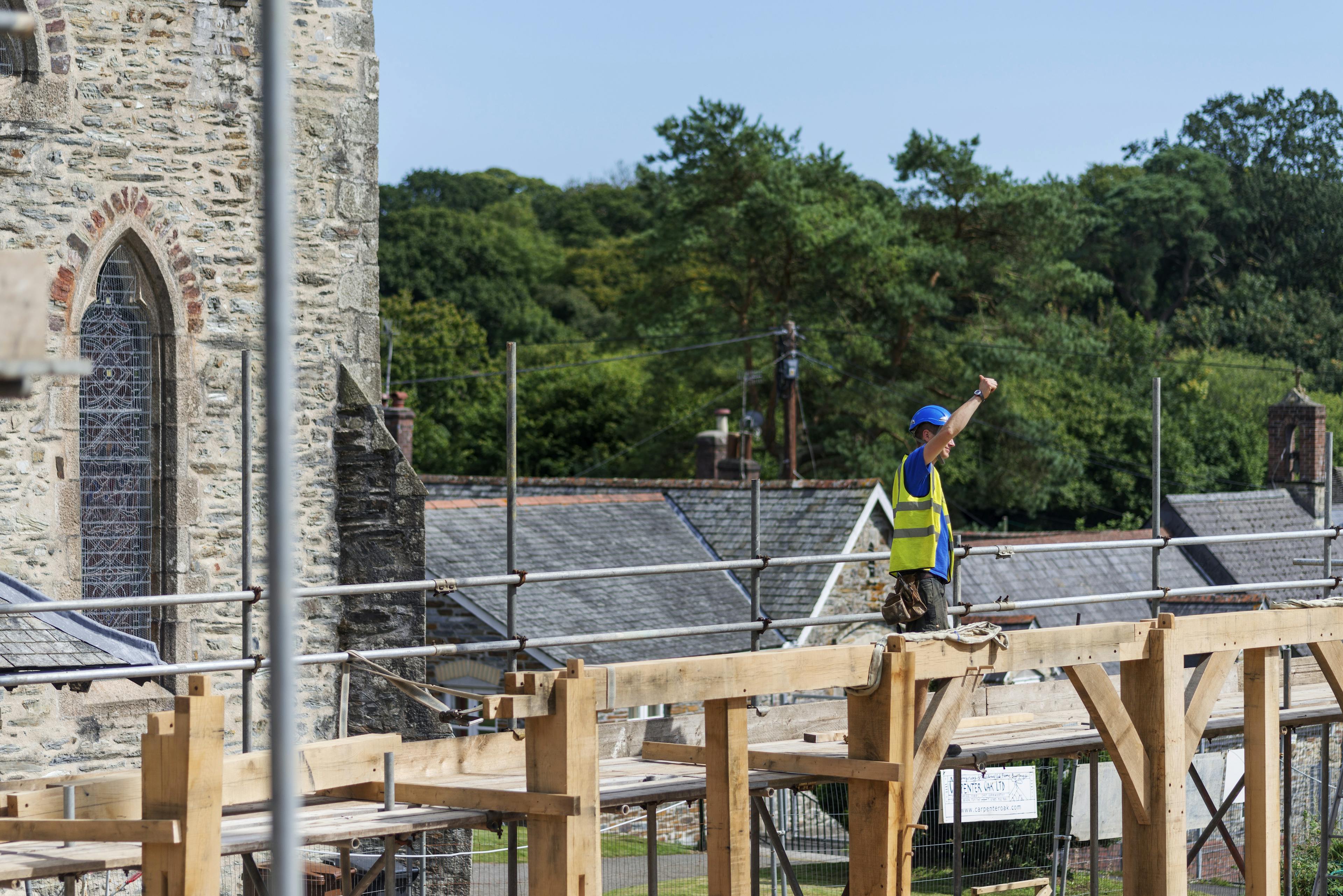 A carpenter on scaffolding next to an oak frame during construction with a church behind