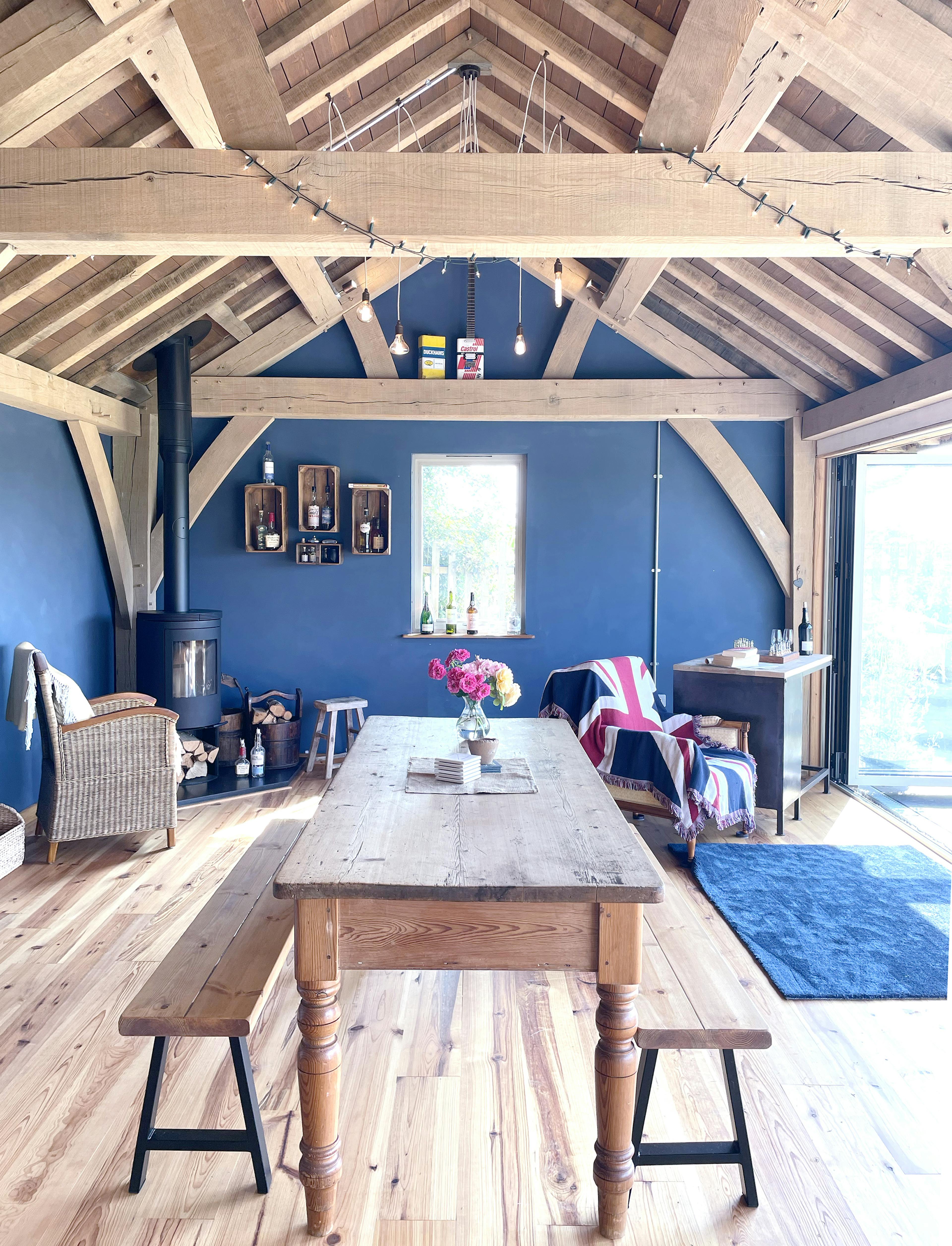 The inside of a 2-bay oak framed extension with a log burner, wooden table and bench, and bi fold doors opening out to a garden