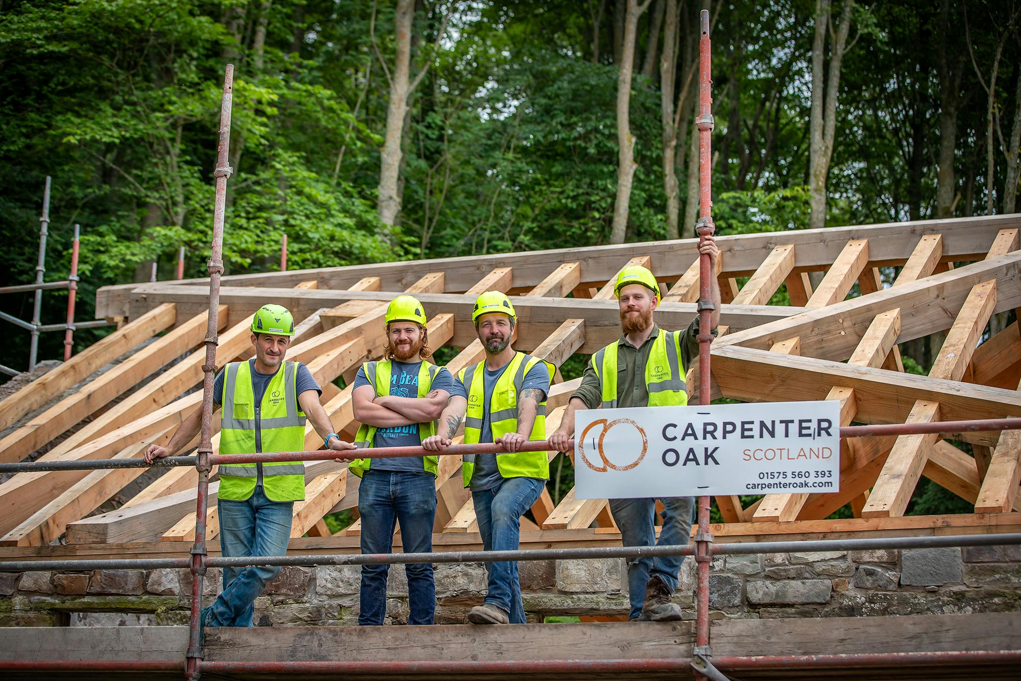 Four carpenters in hard hats and high vis jackets on scaffolding by a timber frame