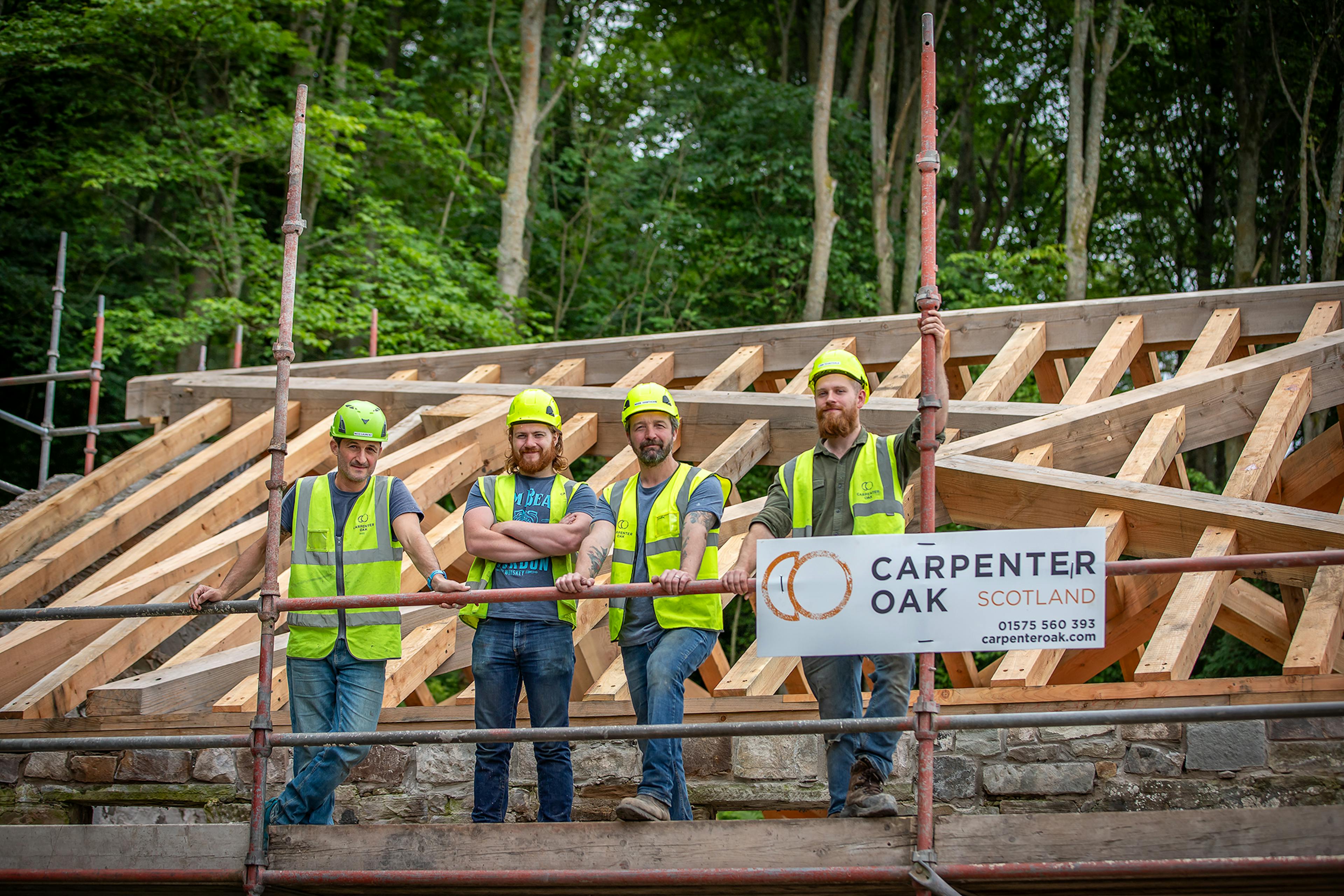 Four carpenters in hard hats and high vis jackets on scaffolding by a timber frame