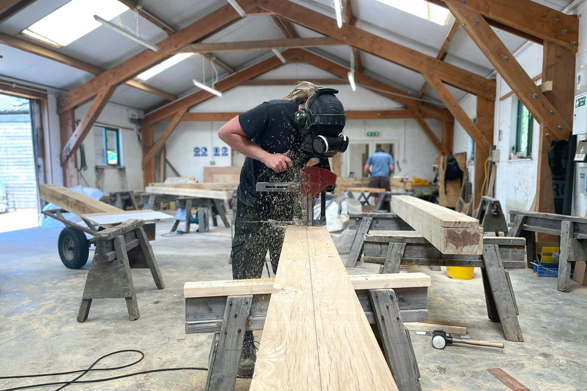 A carpenter in a workshop uses a hand held power tool on an oak frame for a structural timber frame