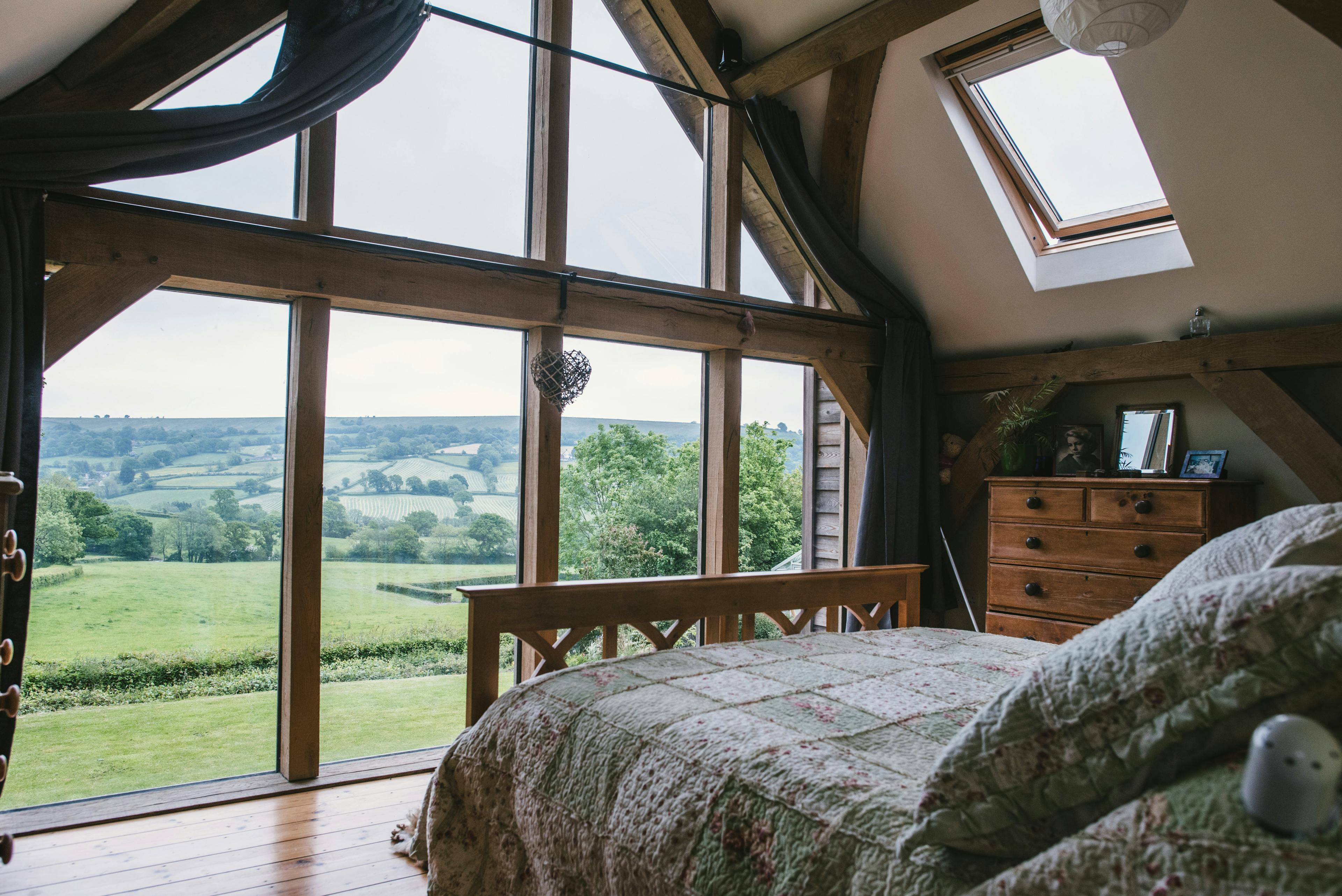 Interior view of an oak-framed bedroom with a glazed gable end, offering views of the countryside