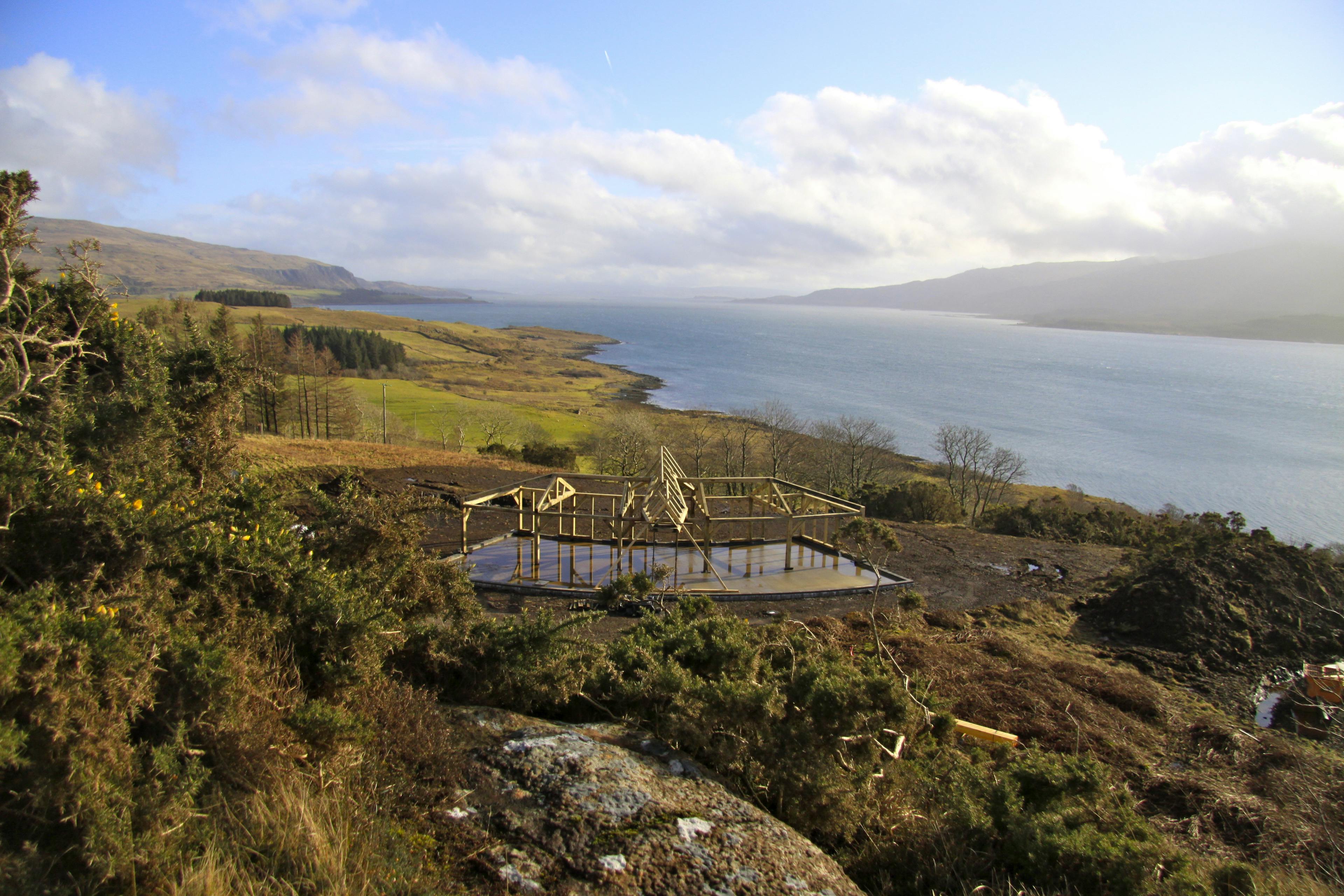Site installation of a unique, curved oak-framed family home with a pitched central roof lantern, overlooking a loch