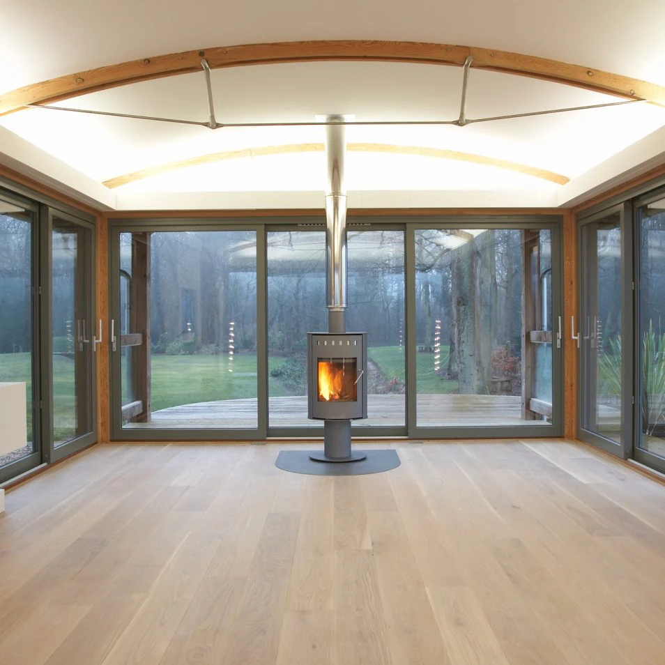Douglas Fir-Framed Lounge Featuring a Central Log Burner, Curved Trusses with Steel Fixings for a Contemporary Touch, and Wrap-Around Glazing Offering Expansive Garden and Countryside Views