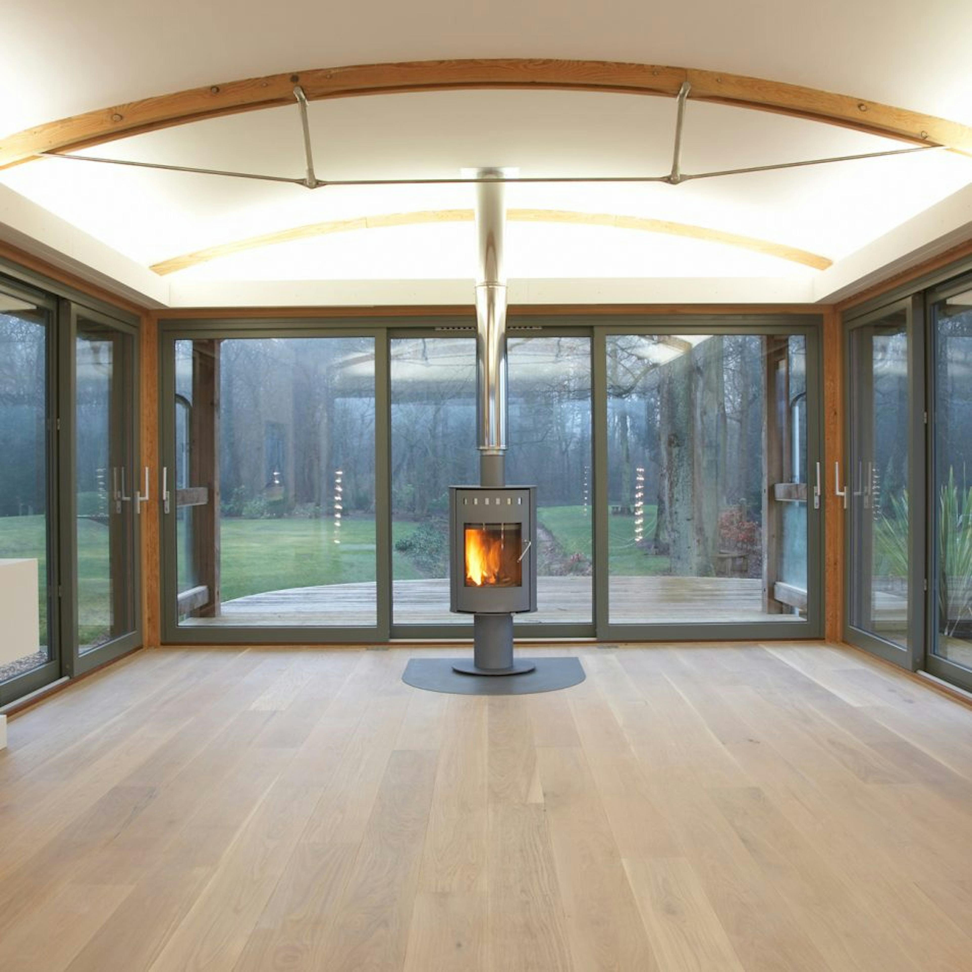 Douglas Fir-Framed Lounge Featuring a Central Log Burner, Curved Trusses with Steel Fixings for a Contemporary Touch, and Wrap-Around Glazing Offering Expansive Garden and Countryside Views