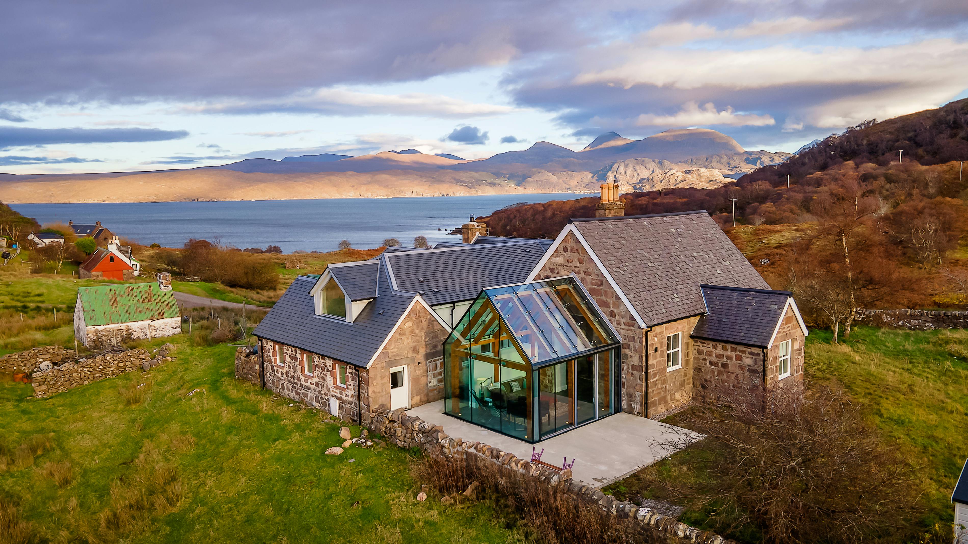 Glazed extension with an internal timber-framed structure attached to a stone cottage, located on the coastline of Scotland, blending modern design with traditional architecture