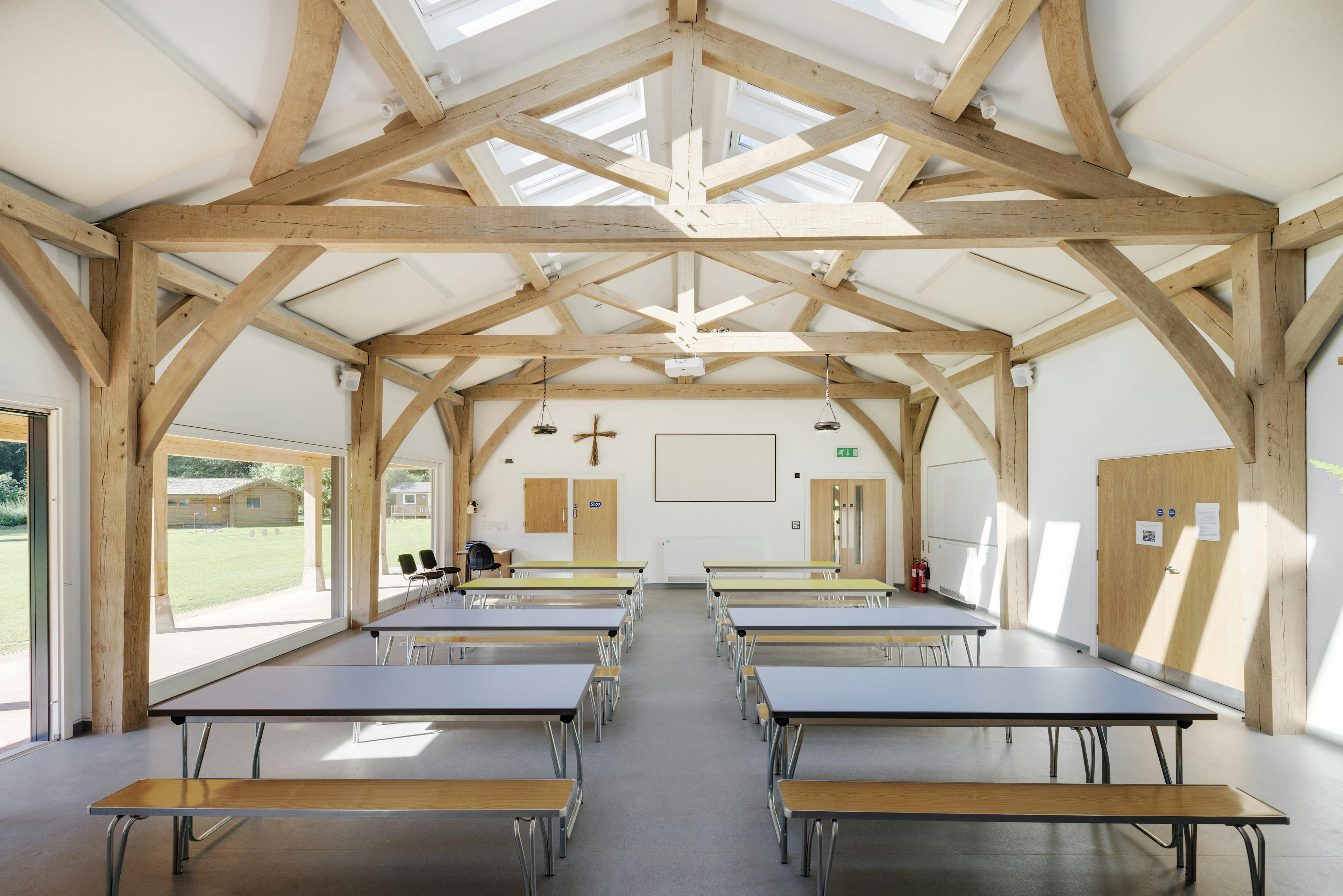 Interior view of the 4-bay multifunctional space at Great Wood Camp, featuring exposed kingpost trusses and roof lanterns that fill the space with natural light