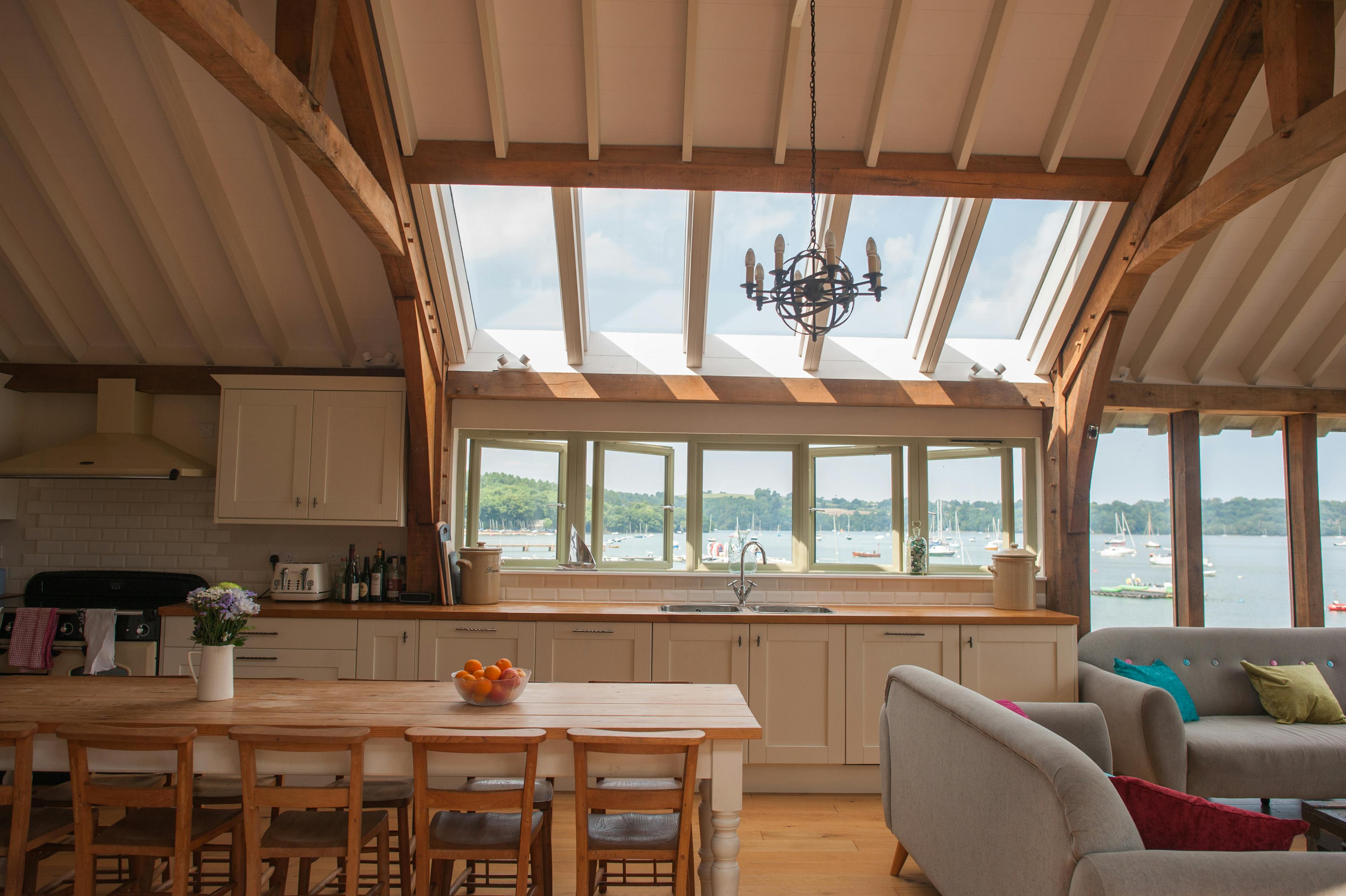 An internal view of an open-plan kitchen and dining room with a vaulted ceiling and roof lanterns, allowing natural light to flood the space while showcasing expansive views of the estuary