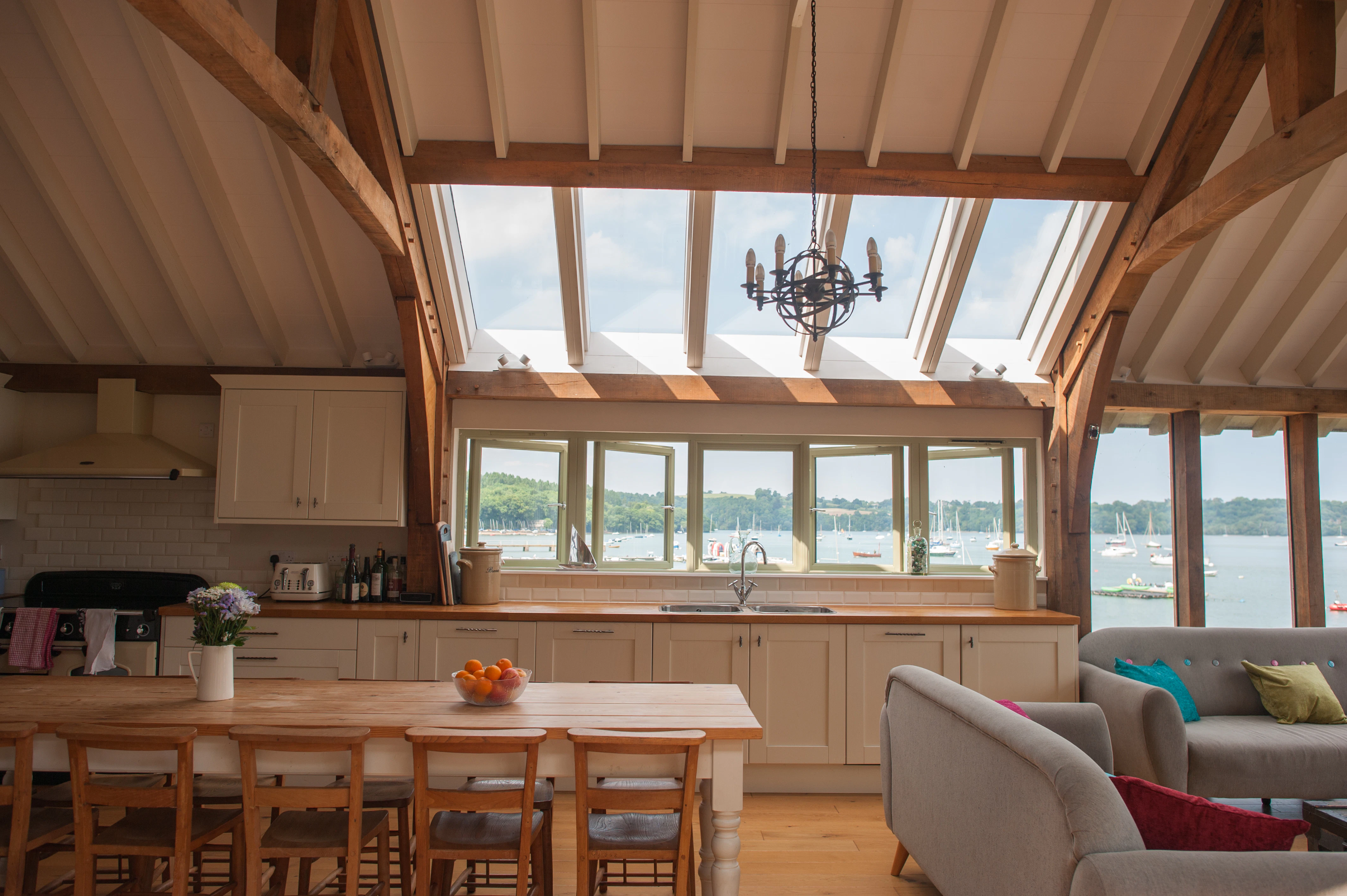 An internal view of an open-plan kitchen and dining room with a vaulted ceiling and roof lanterns, allowing natural light to flood the space while showcasing expansive views of the estuary