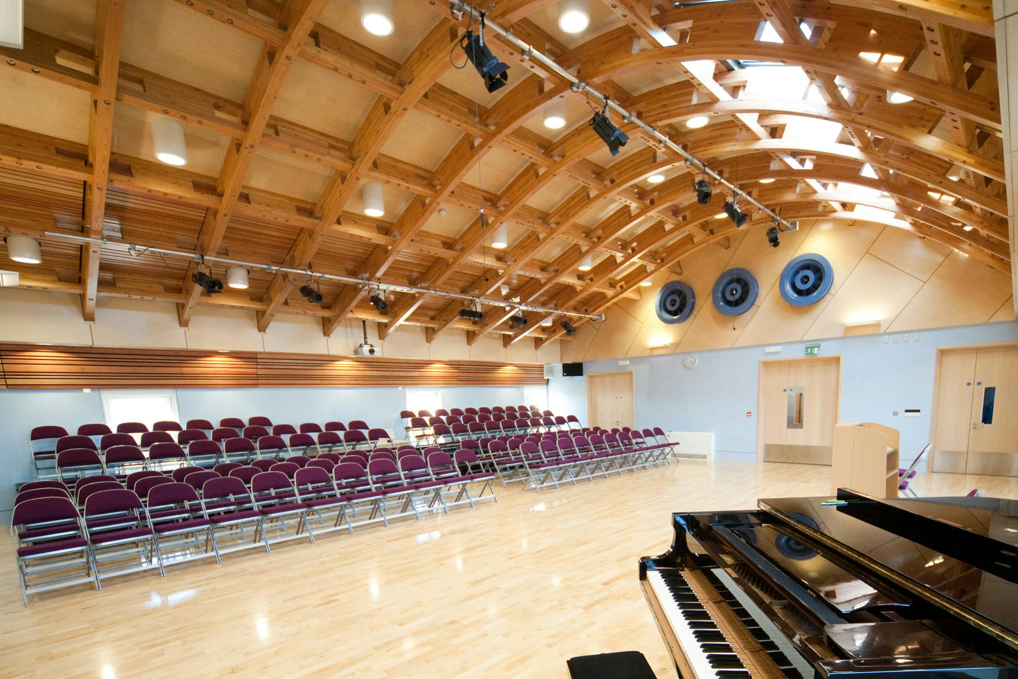 A hall with a glulam gridshell roof structure and rows of seating for an audience