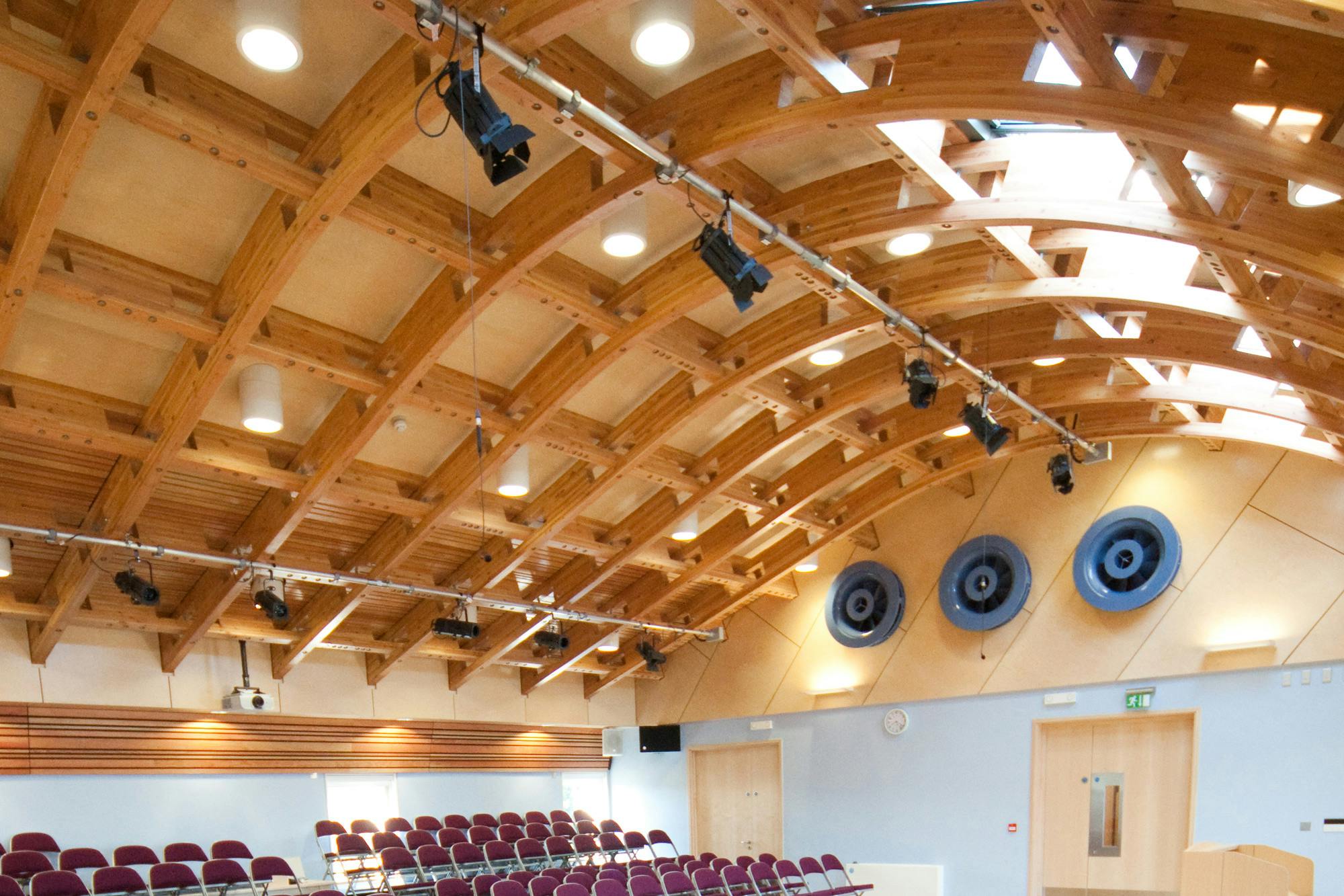 A hall with a glulam gridshell roof structure and rows of seating for an audience