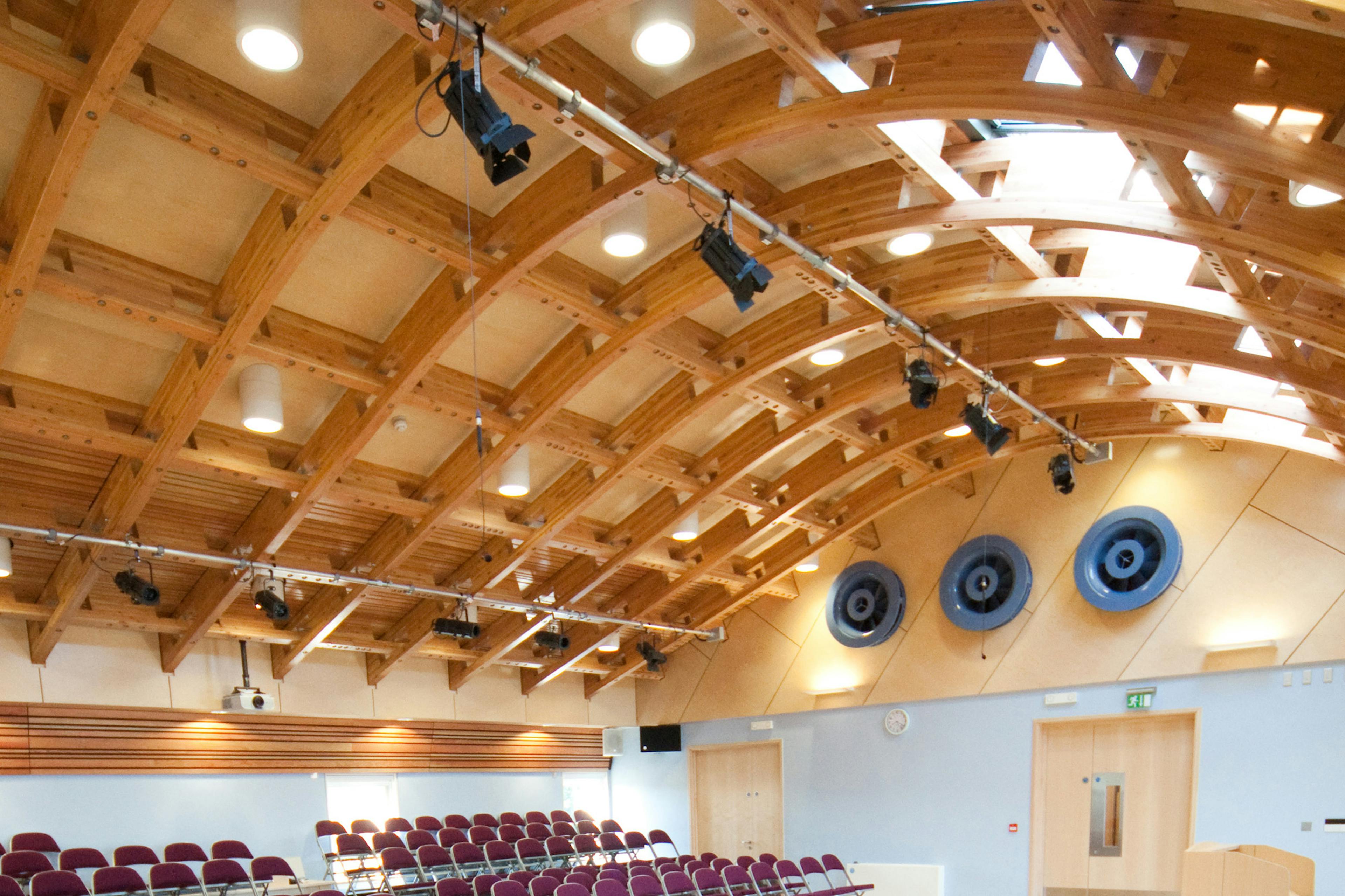 A hall with a glulam gridshell roof structure and rows of seating for an audience