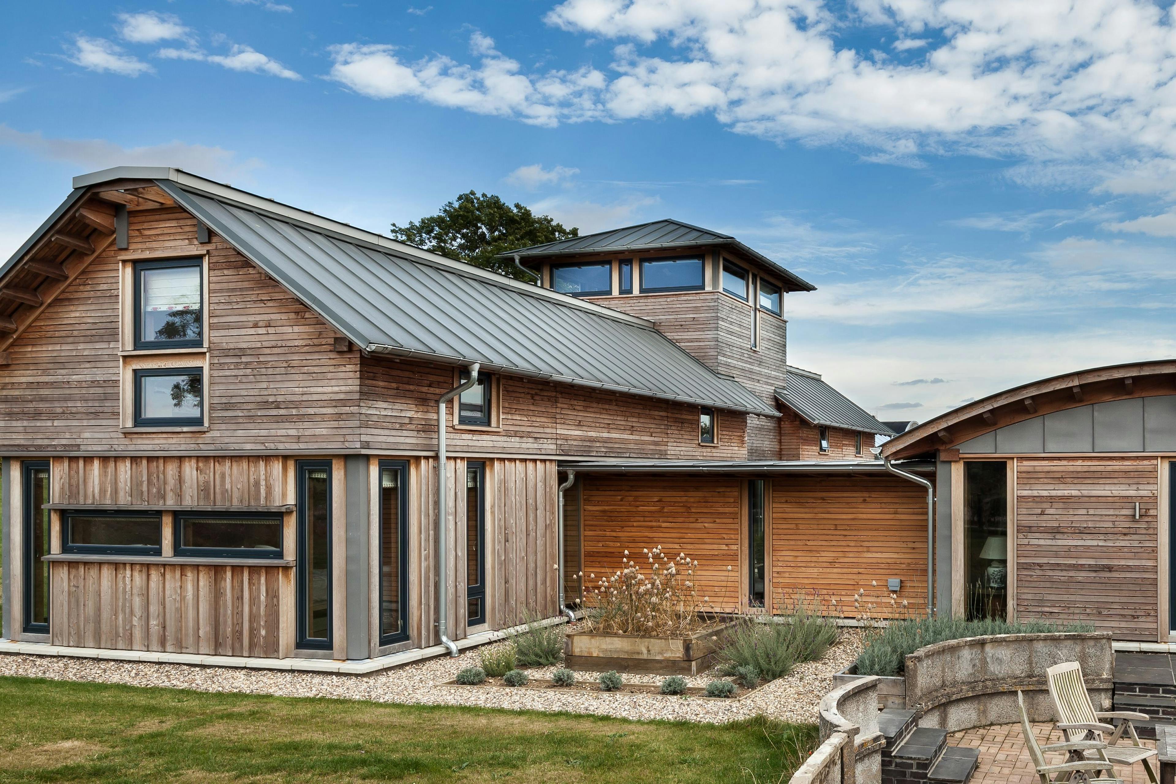 The rear of a timber clad large family farmhouse with a tower surrounded by a green lawn and with a patio area and outdoor seating