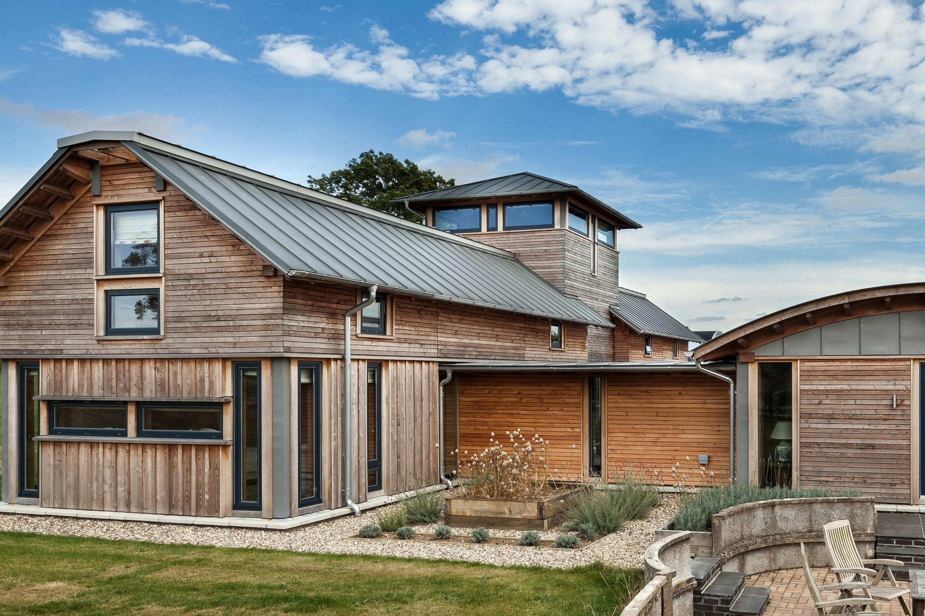 The rear of a timber clad large family farmhouse with a tower surrounded by a green lawn and with a patio area and outdoor seating