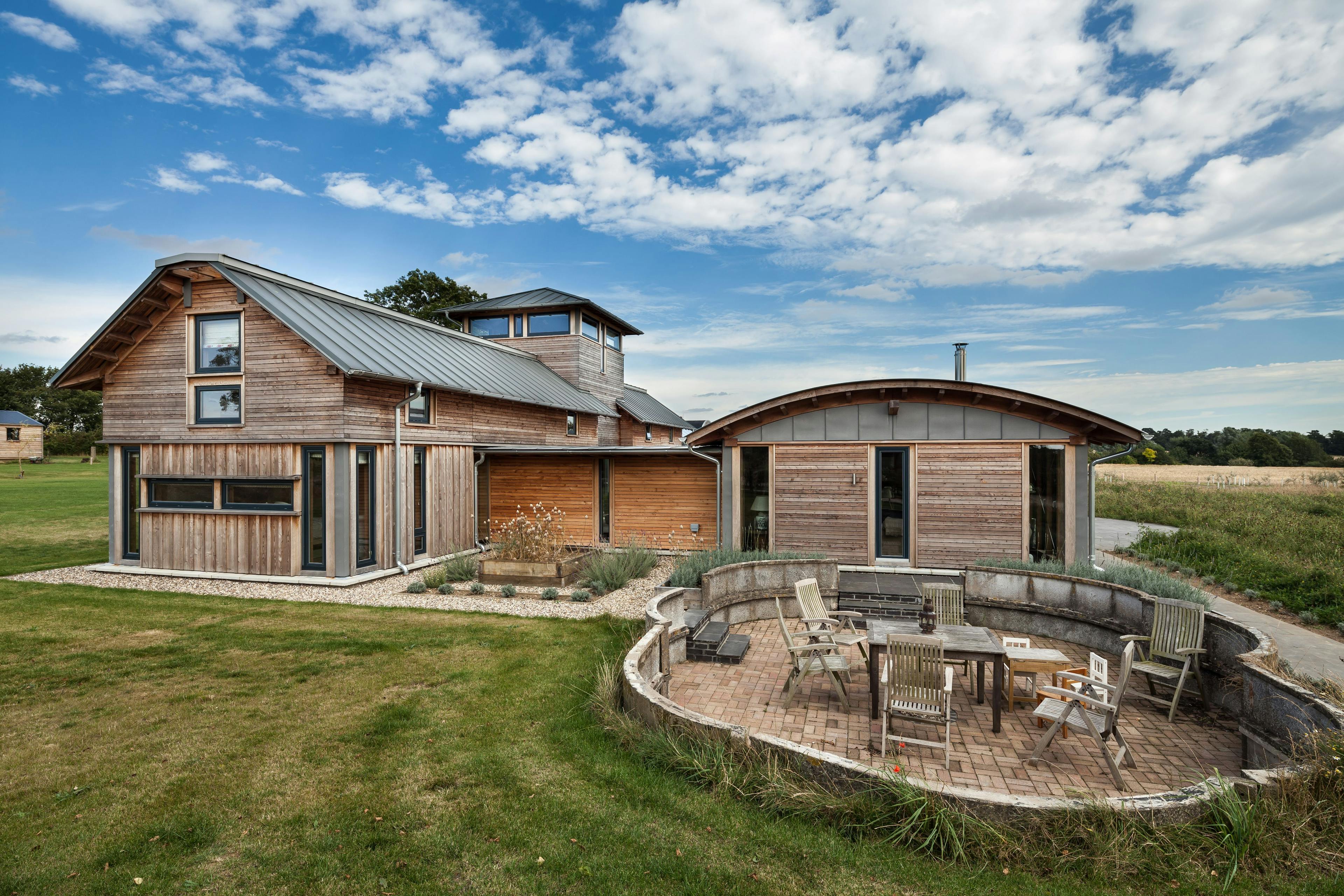 The rear of a timber clad large family farmhouse with a tower surrounded by a green lawn and with a patio area and outdoor seating