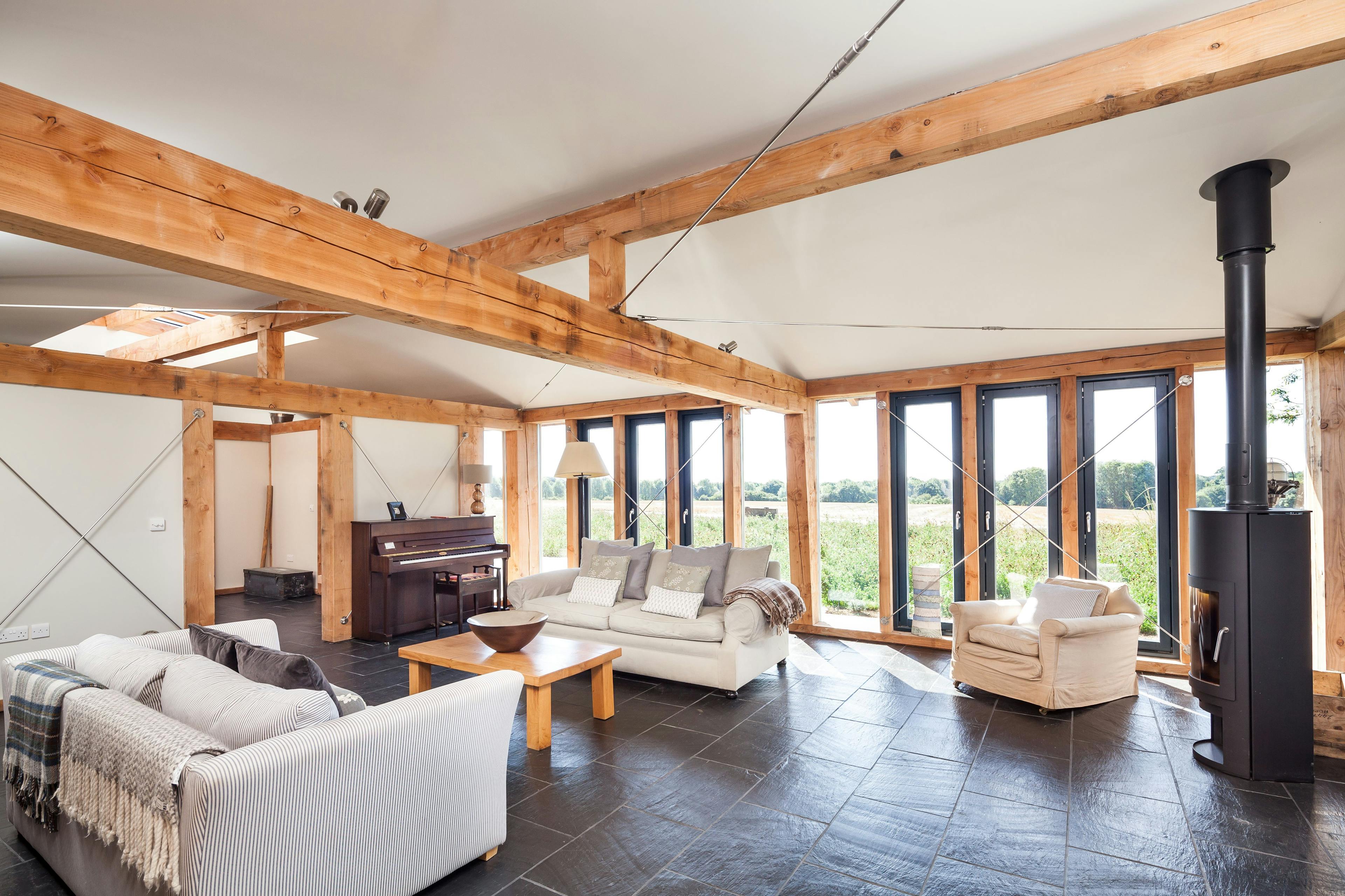 The sitting area in a Douglas fir framed family farmhouse with sofas, piano and log burner and large glazing with views of the garden