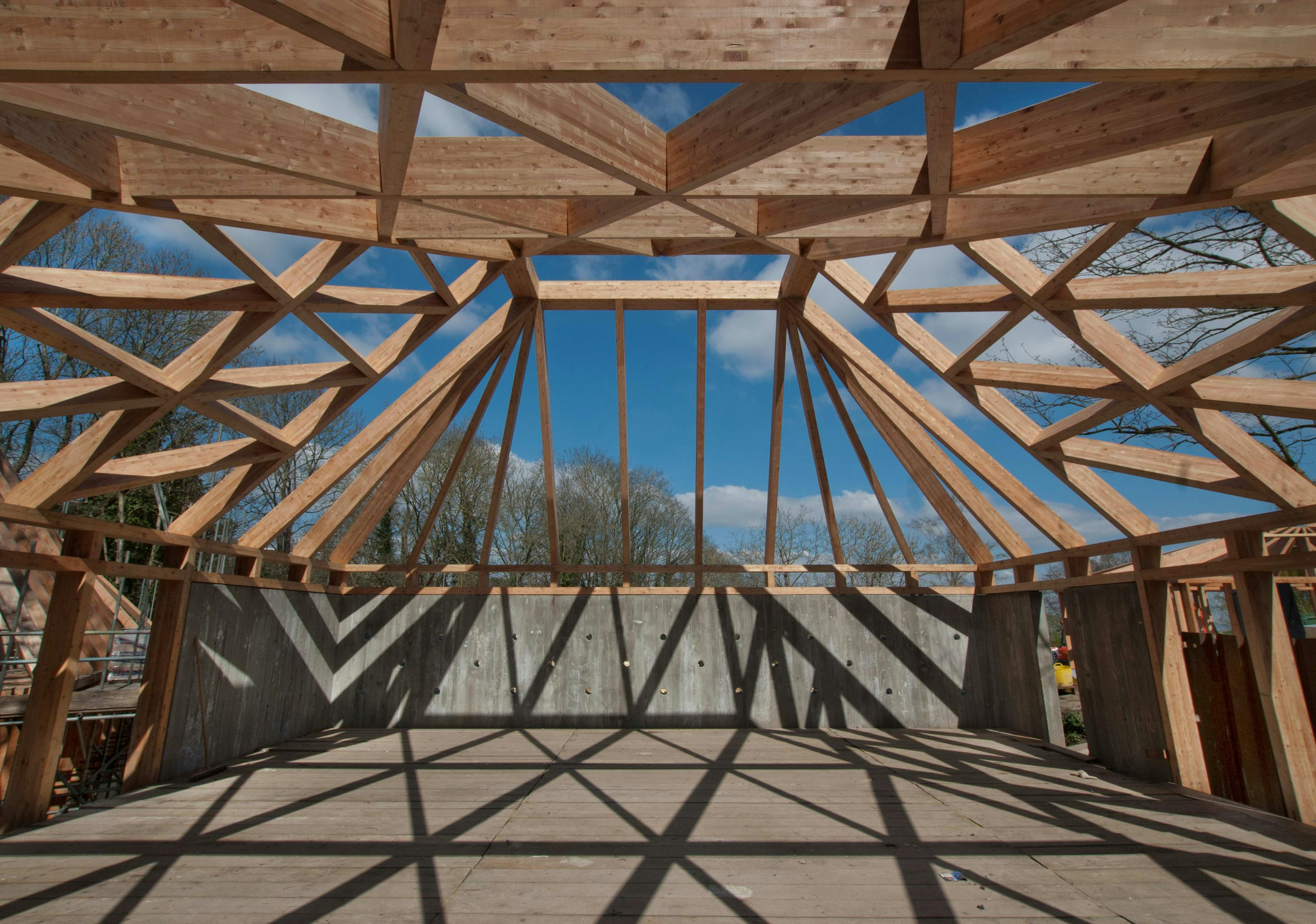 The sun shines through a larch glulam frame of a pyramidic building causing shadows on a construction site during installation