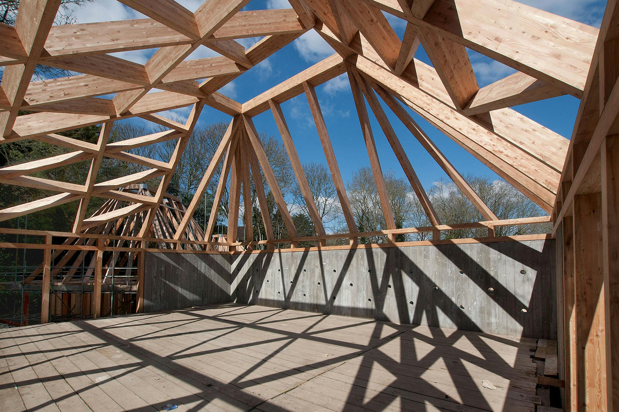 The sun shines through a larch glulam frame of a pyramidic building causing shadows on a construction site during installation