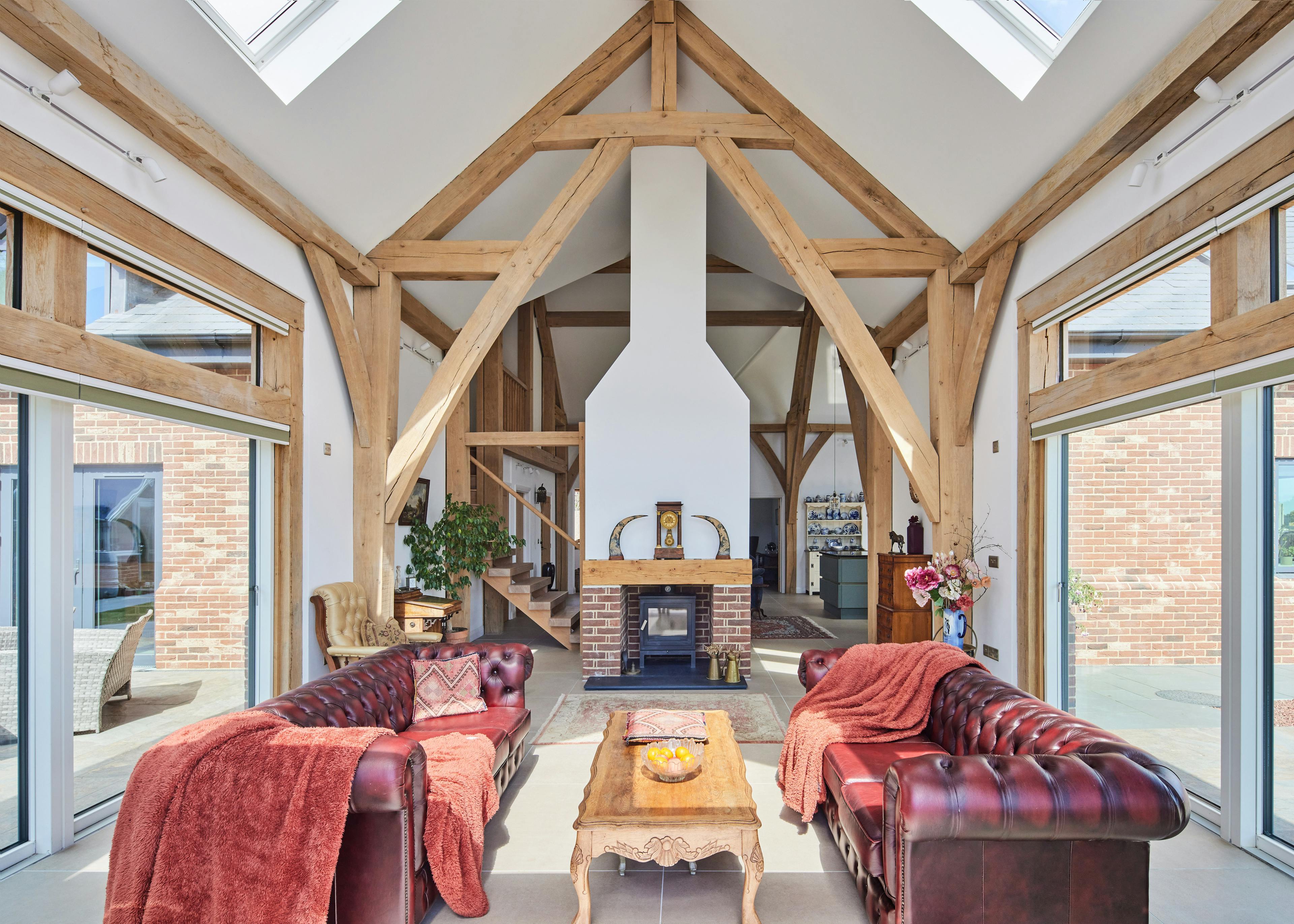 An open plan living sitting area with red leather sofas and log burner in an oak framed home with a double height vaulted ceiling.