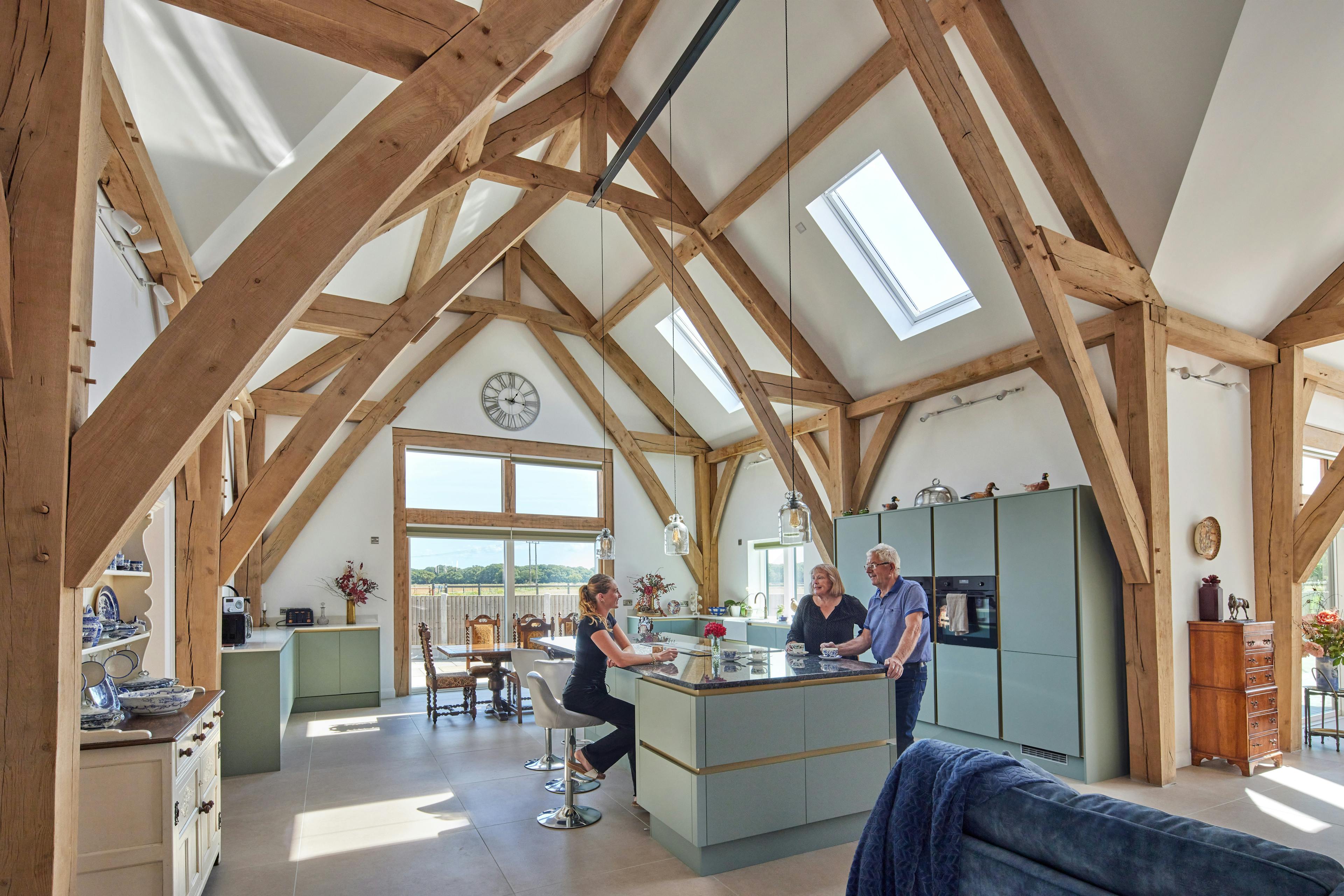 People sitting at a kitchen island in an open plan dining and kitchen area in an oak framed home with a double height vaulted ceiling.