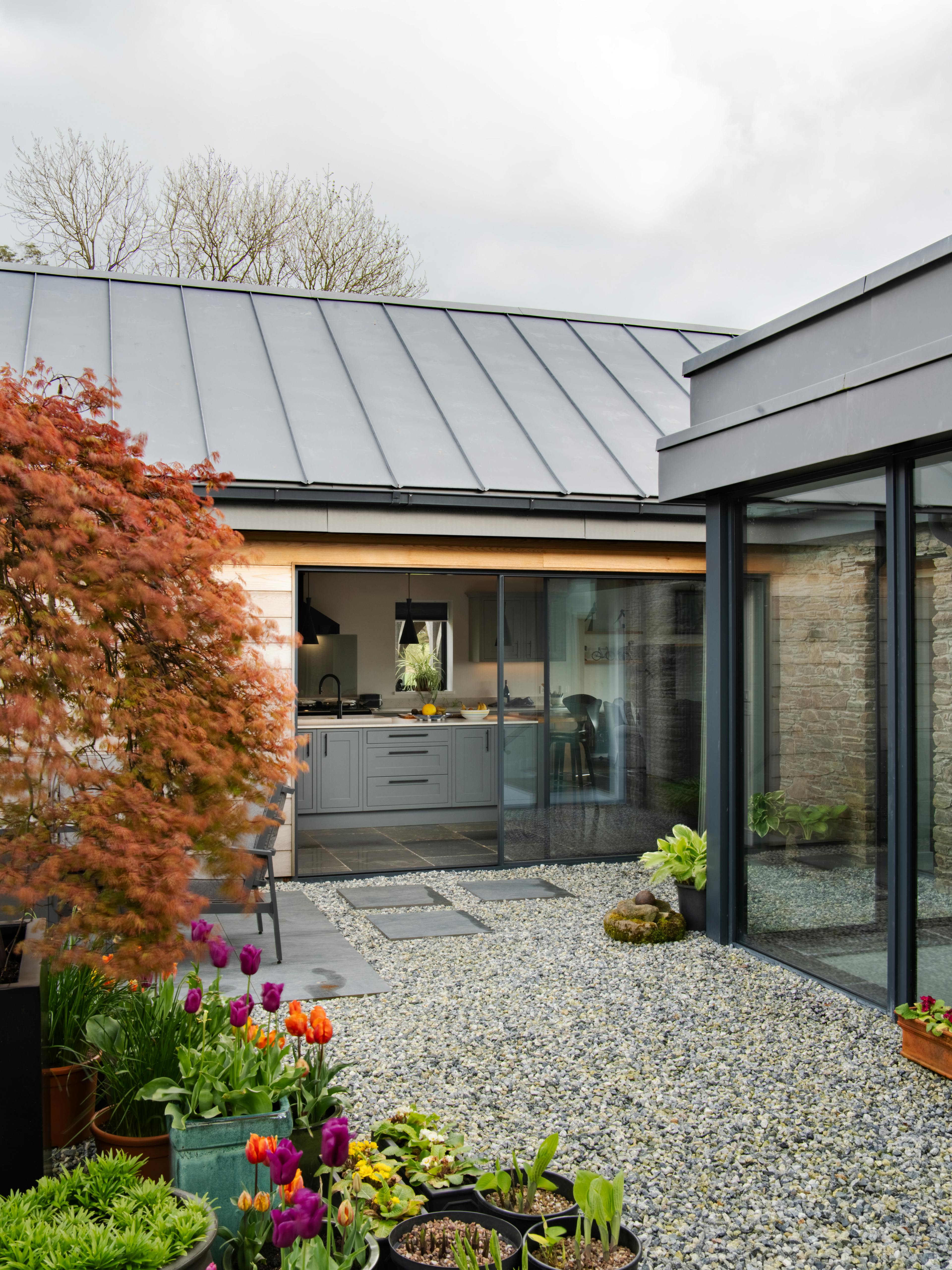 A courtyard with flowers and trees of a stone clad oak framed home with a zinc roof