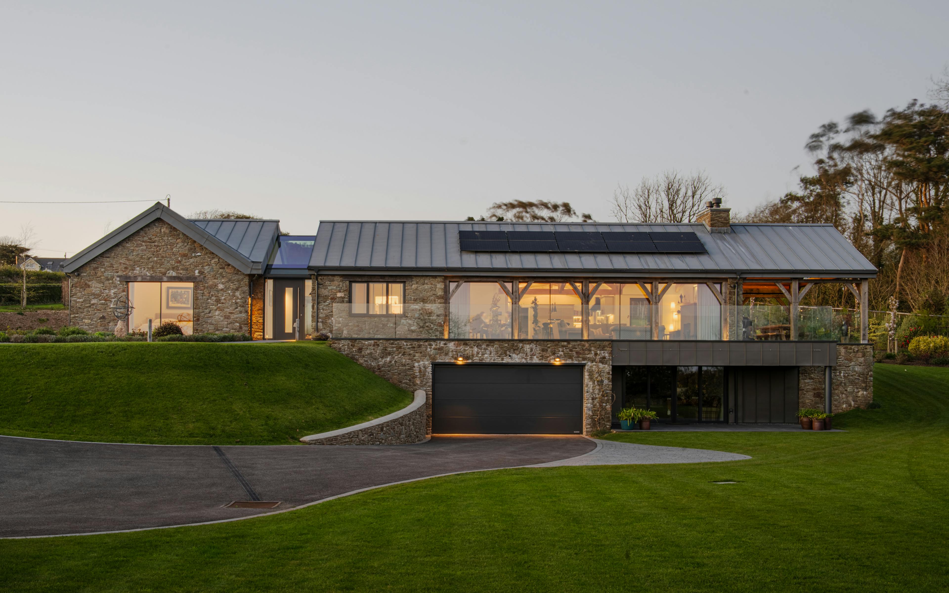 The front of a stone clad oak framed home with a zinc roof, green lawn, lower floor garage and driveway at dusk