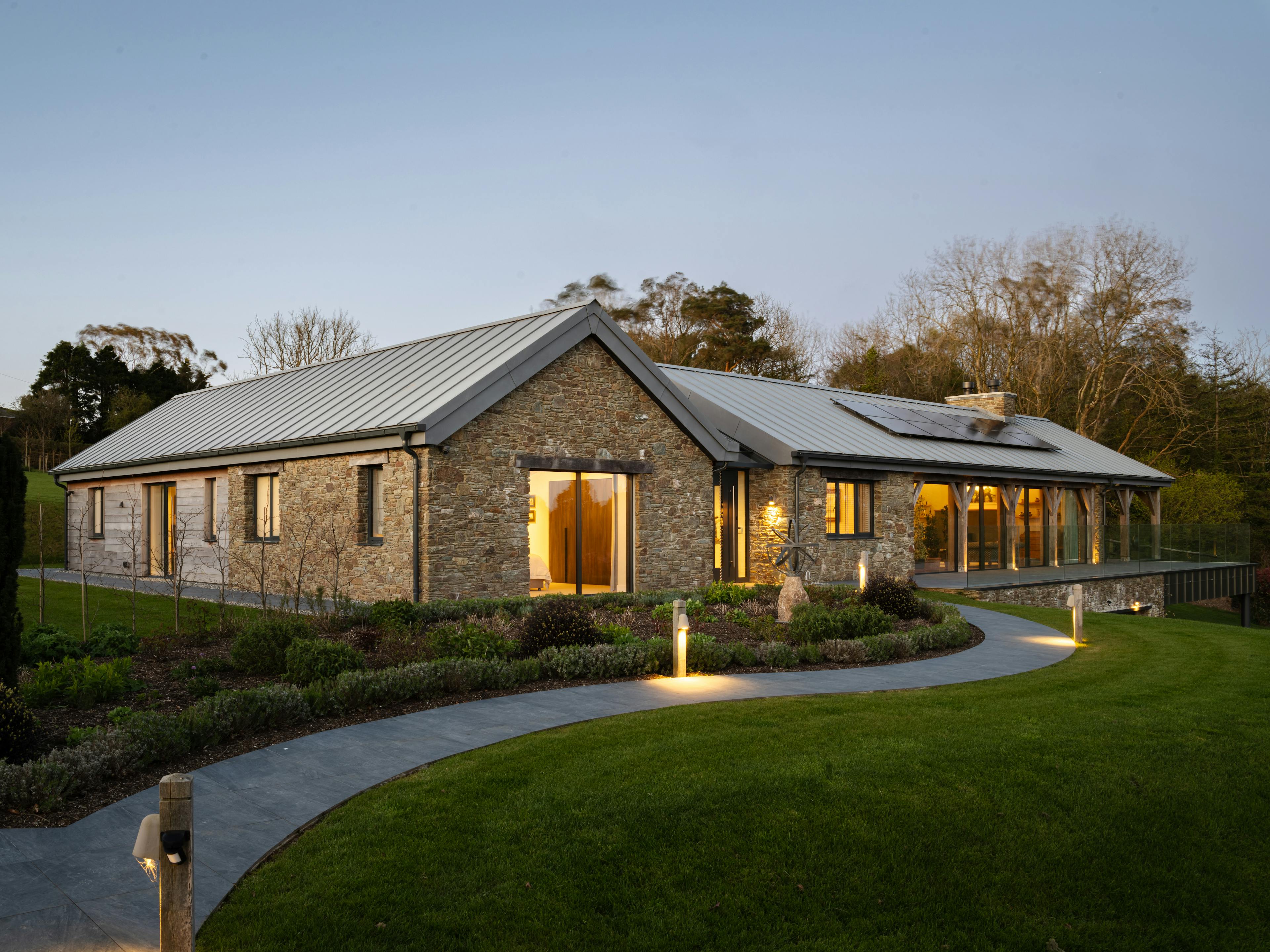The front of a stone clad oak framed home with a zinc roof, green lawn and driveway at dusk
