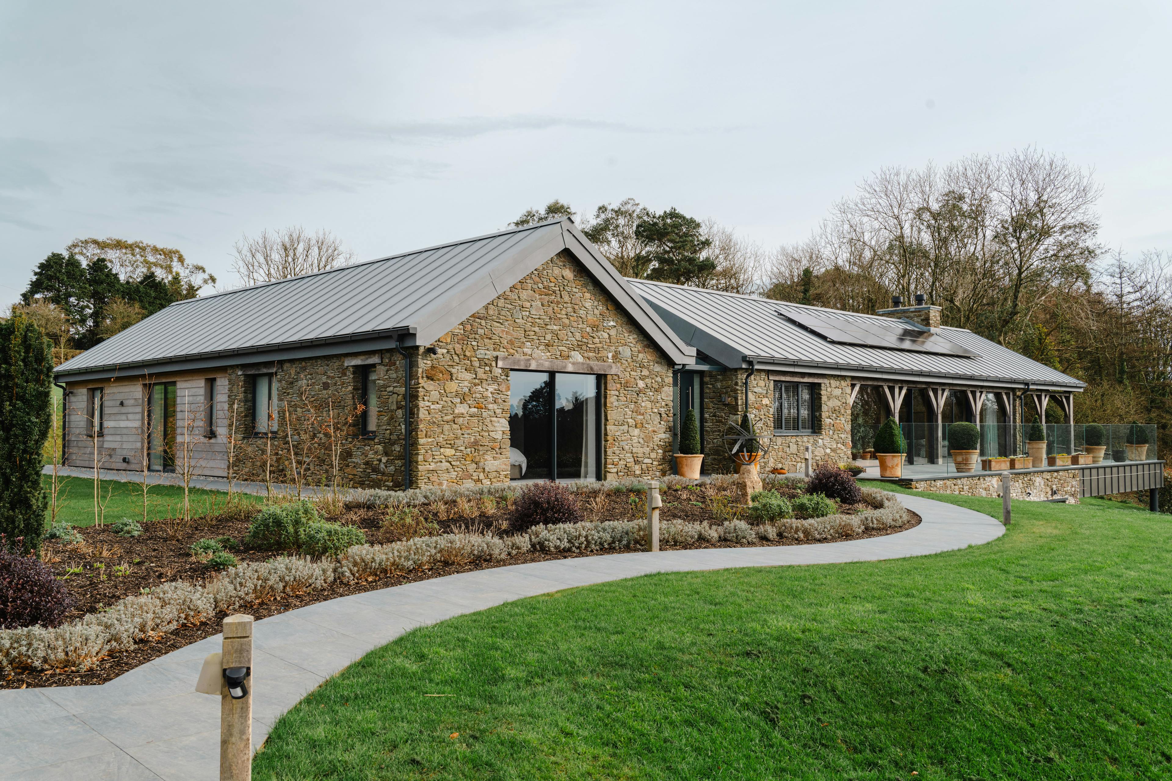 The front of a stone clad oak framed home with a zinc roof, green lawn and driveway