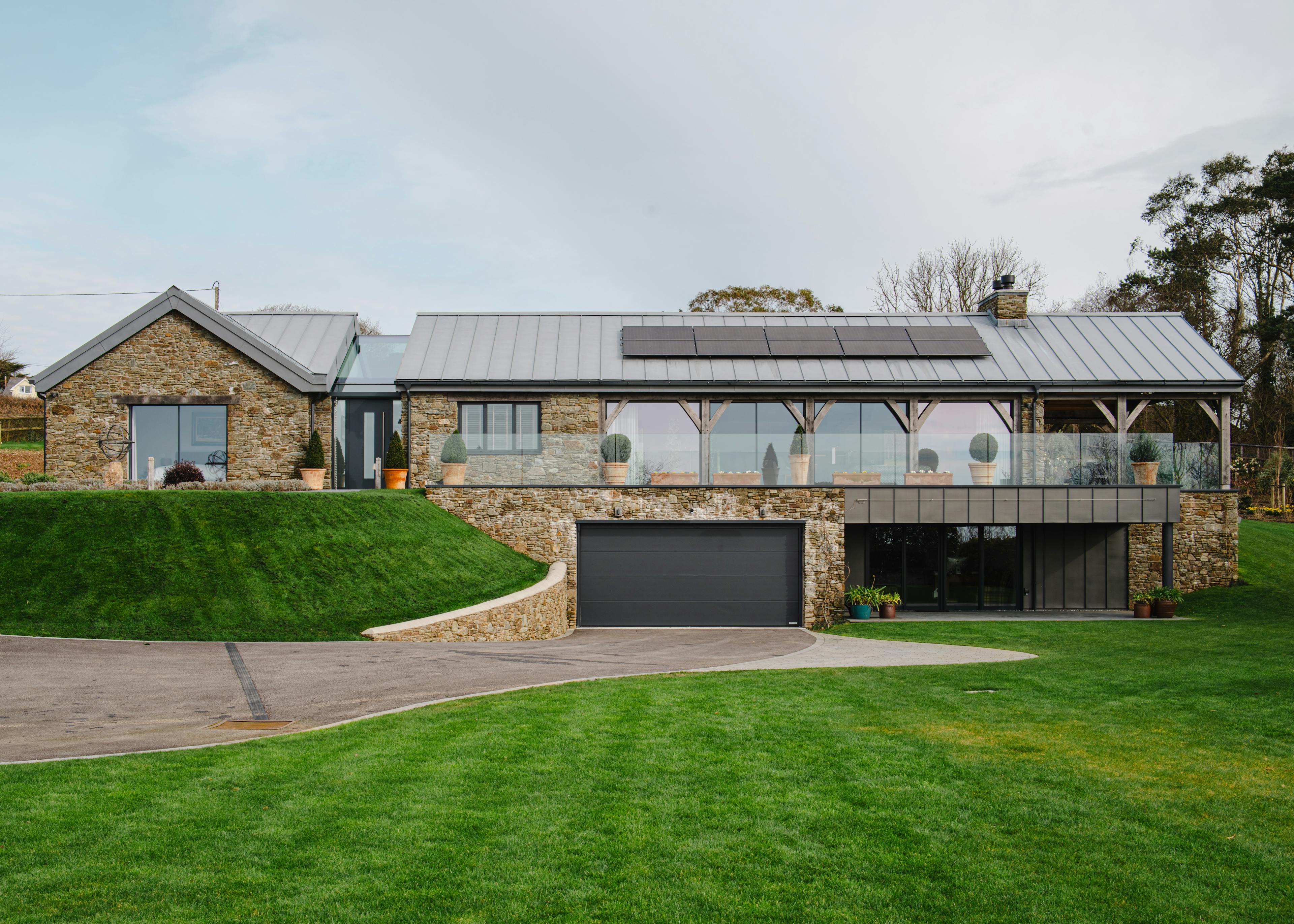 The front of a stone clad oak framed home with a zinc roof, green lawn and driveway 