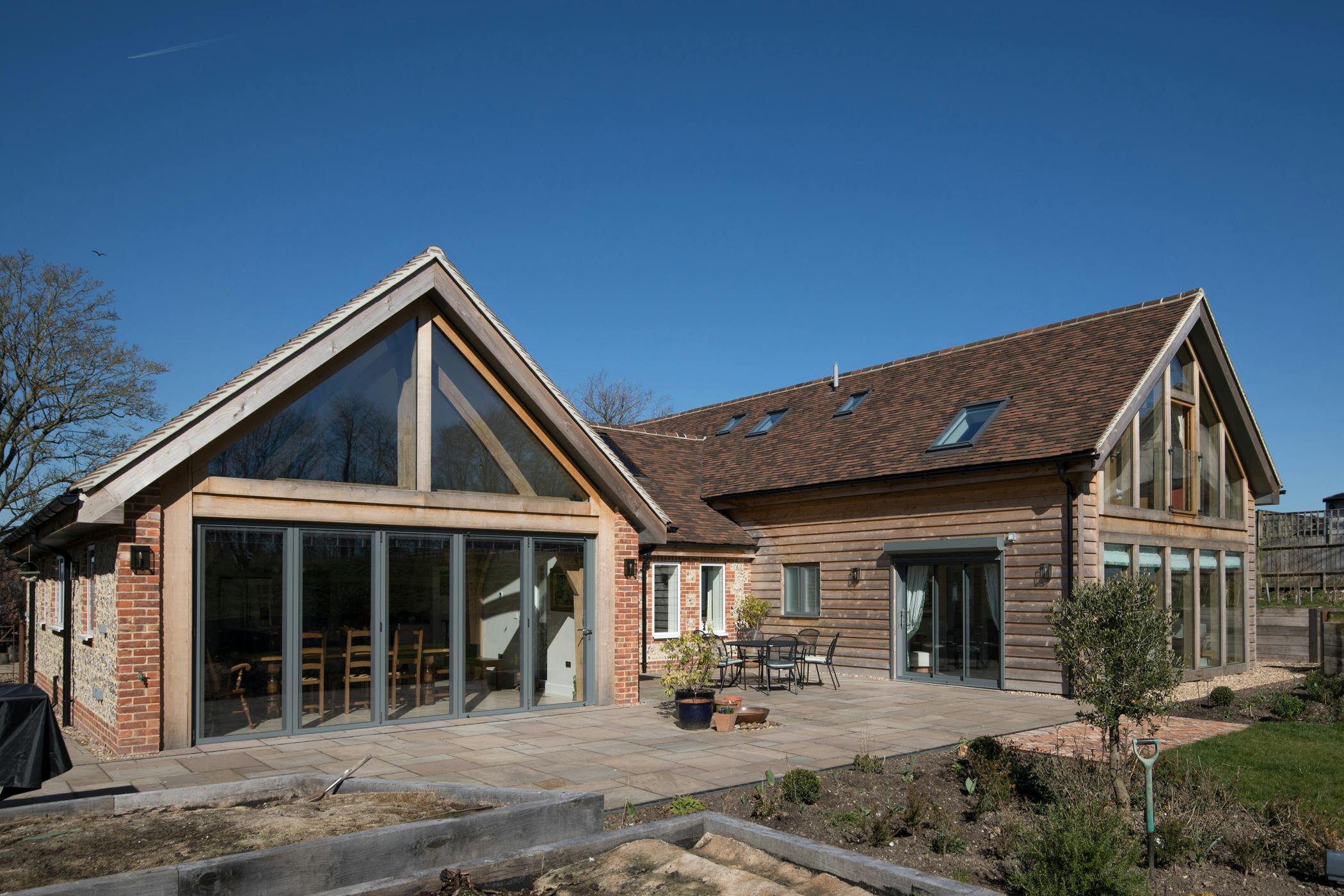 Exterior image of an oak framed house with glazed sliding doors, a sunny patio, and finished in timber cladding and stone and brick work