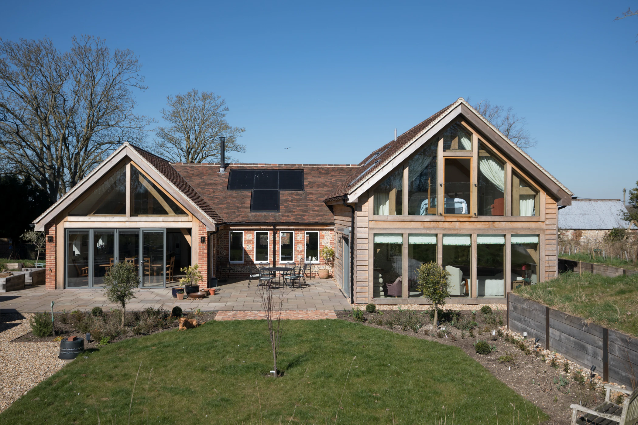 Exterior image of an oak framed house with a sunny patio and green lawn, and finished in timber cladding and stone and brick work
