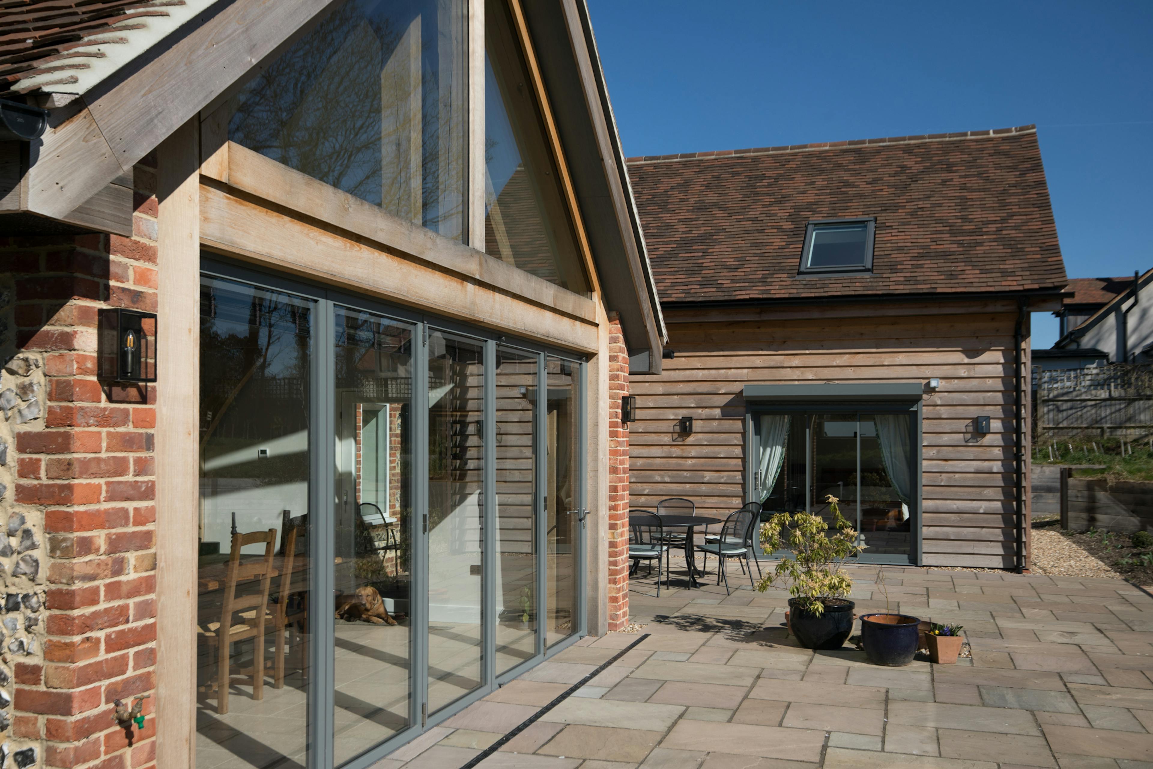 Exterior image of an oak framed house with glazed sliding doors, a sunny patio, and finished in timber cladding and stone and brick work