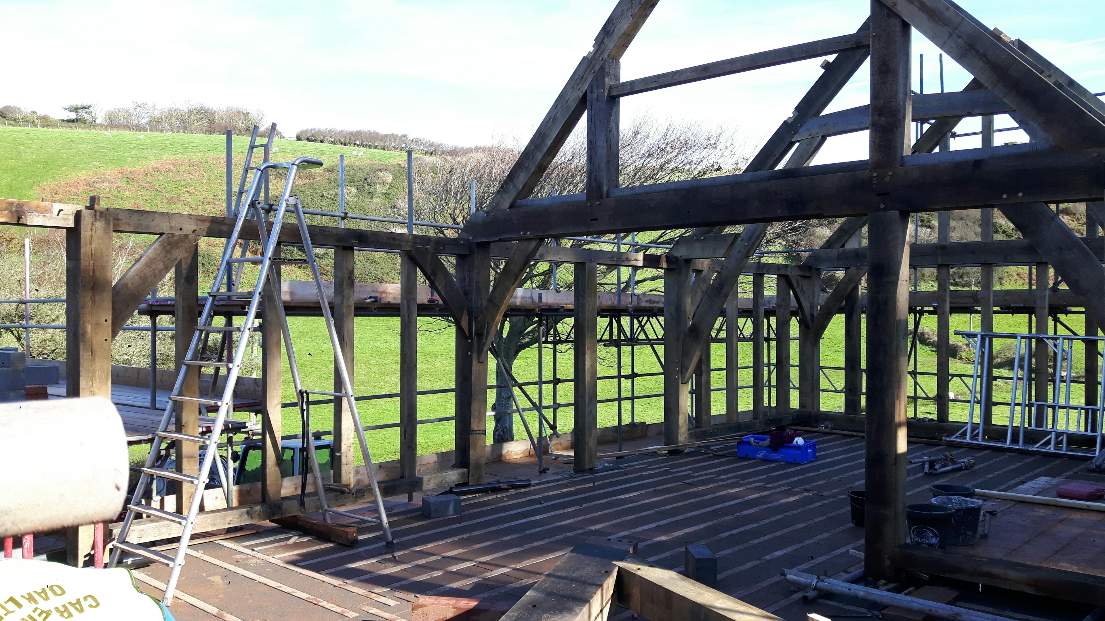 An oak frame being installed on a building site with green fields behind and a blue sky