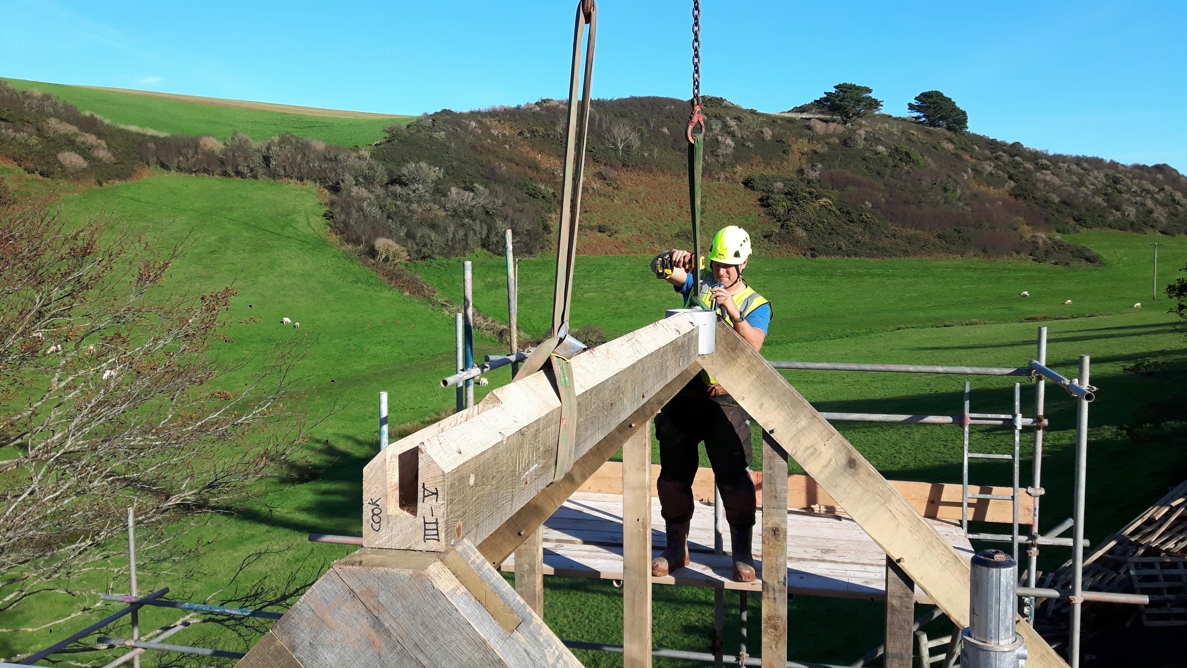 An oak frame being installed by a carpenter on a building site with green fields behind and a blue sky