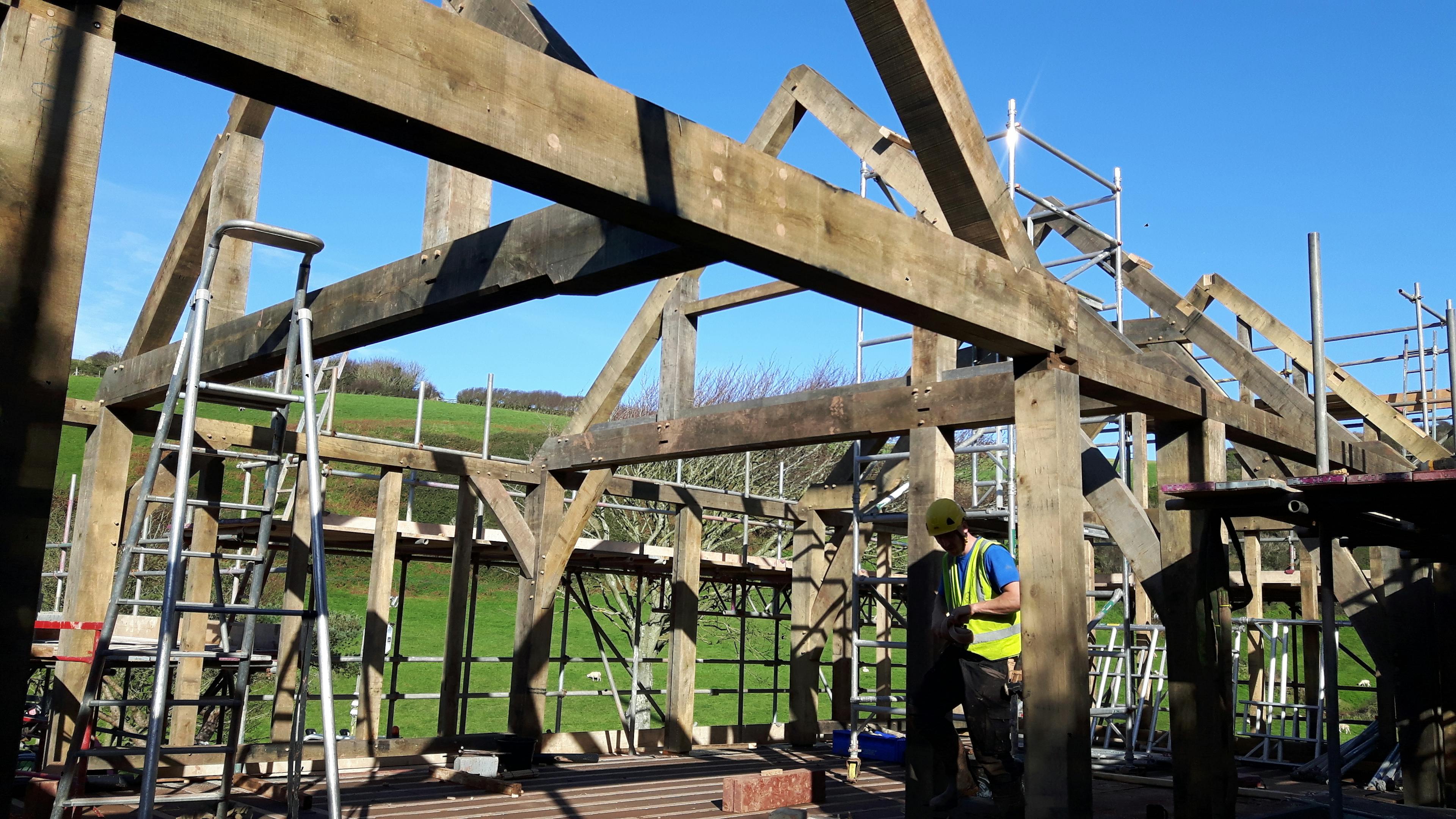 An oak frame being installed on a building site with green fields behind and a blue sky