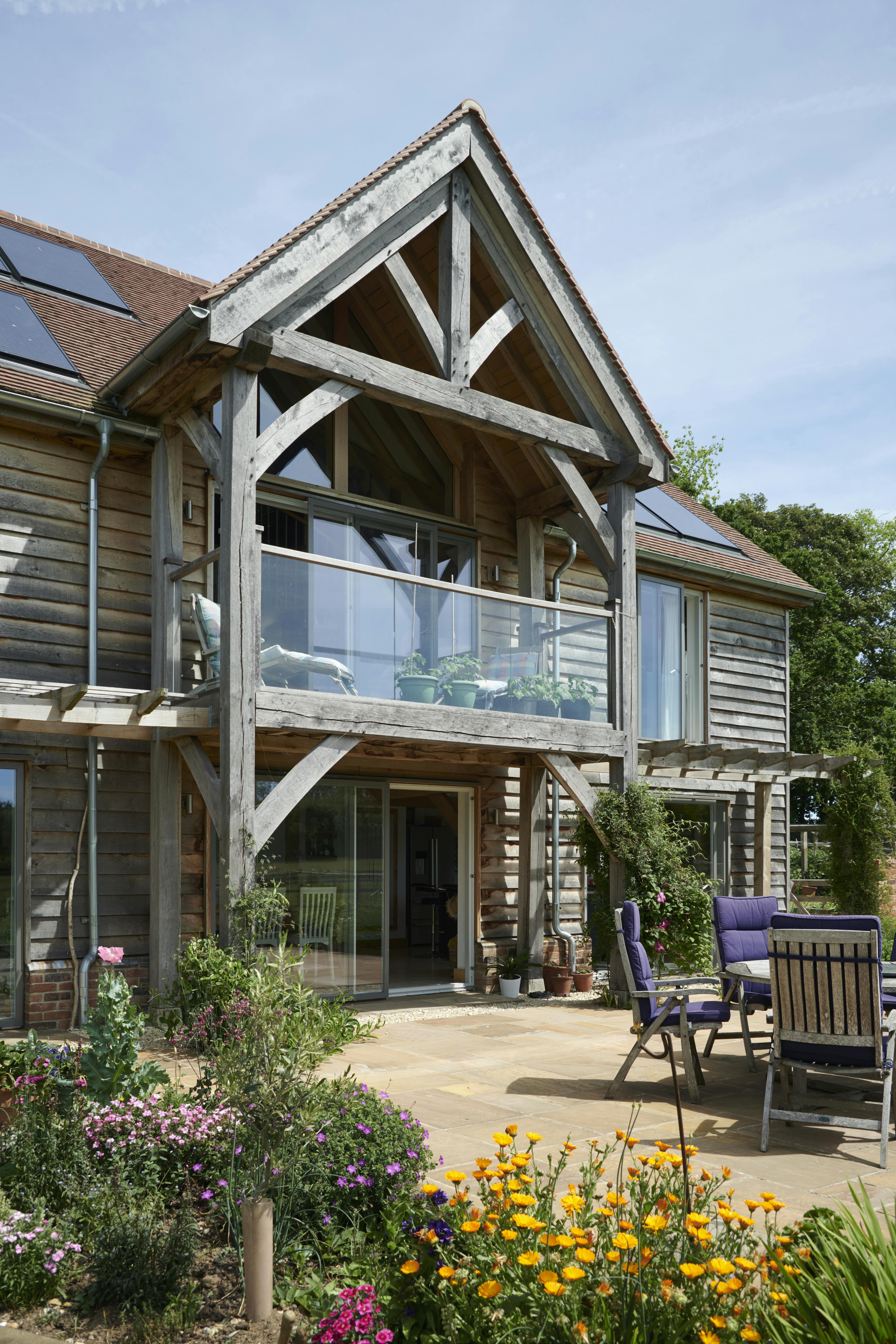Timber clad large oak framed home with an oak balcony, showing back garden, flowers and outdoor table and chairs