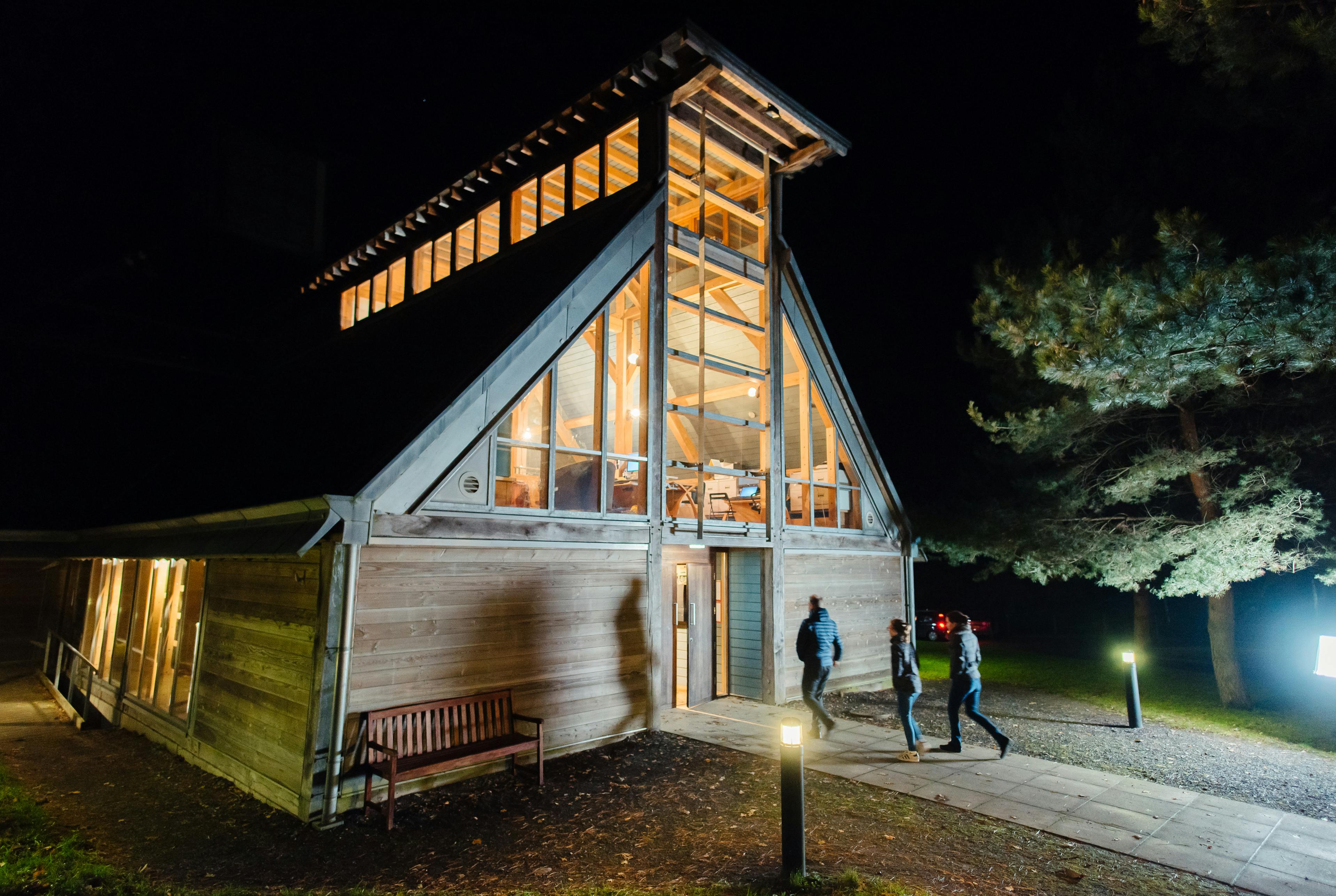 An oak frame theatre with a pyramidal roof lantern, shown at night with lights on an people entering the building
