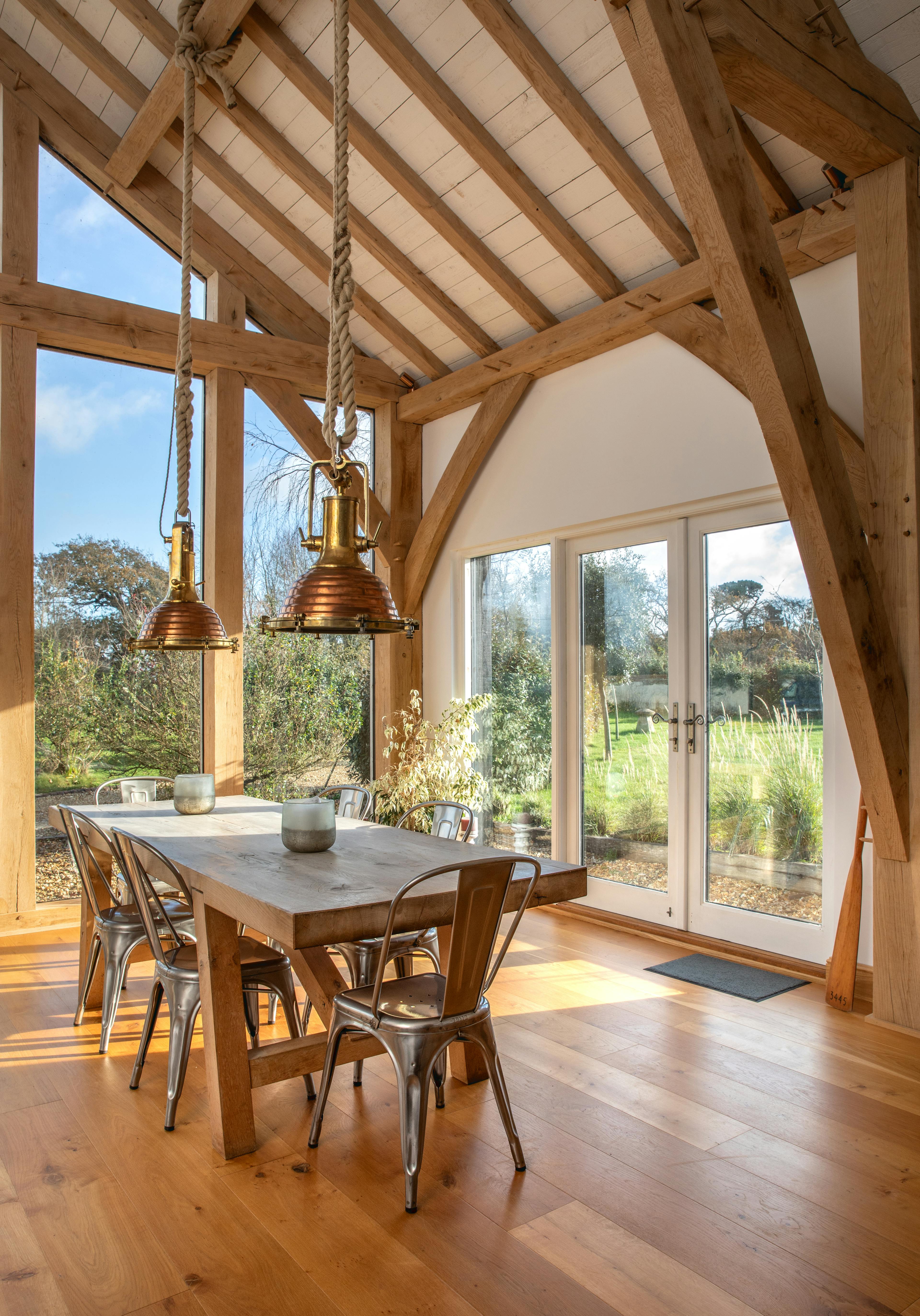 Sun beams shine through the windows on a wooden table and chairs in an oak framed extension with a glazed gable