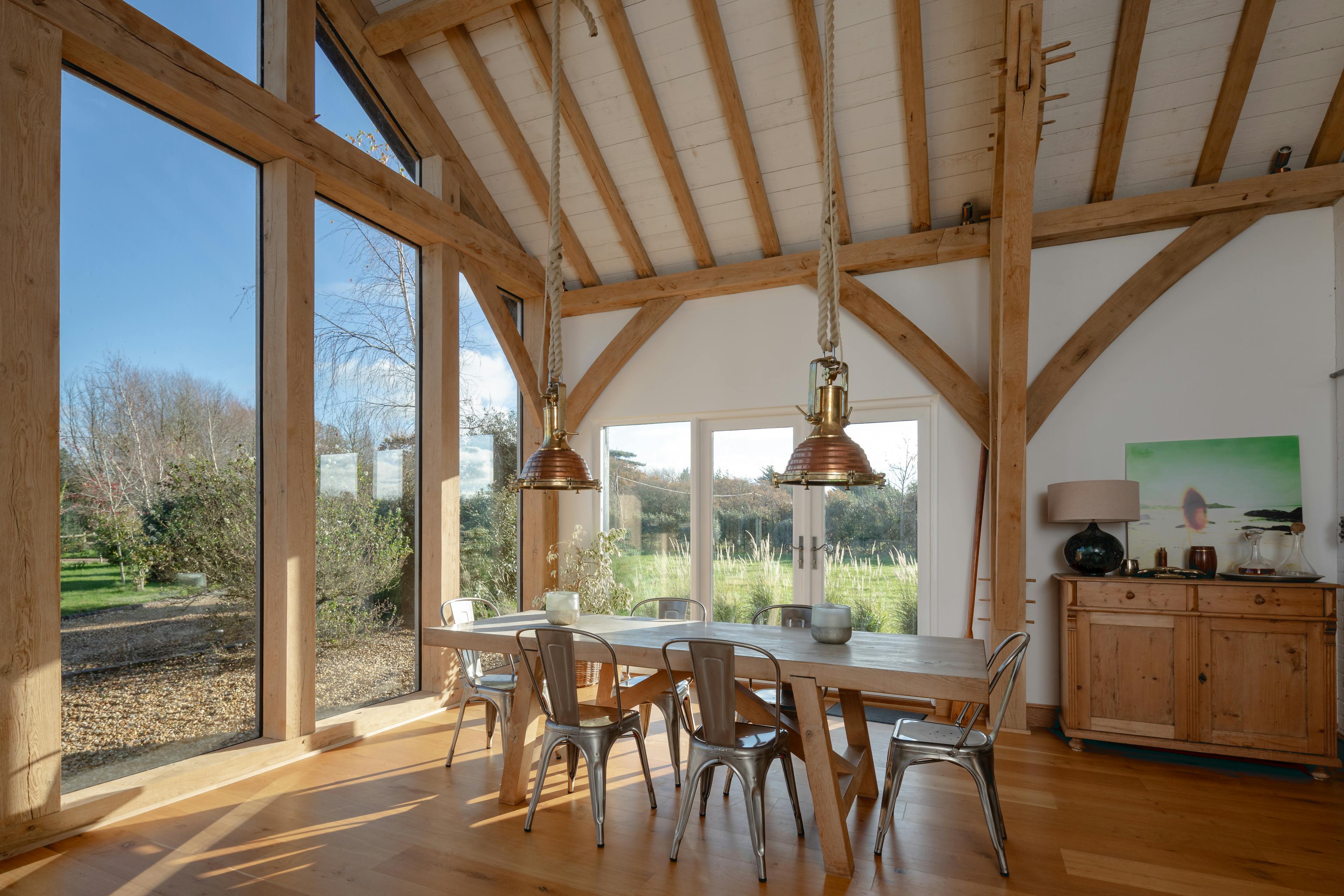 Sun beams shine through the windows on a wooden table and chairs and sideboard in an oak framed extension with a glazed gable