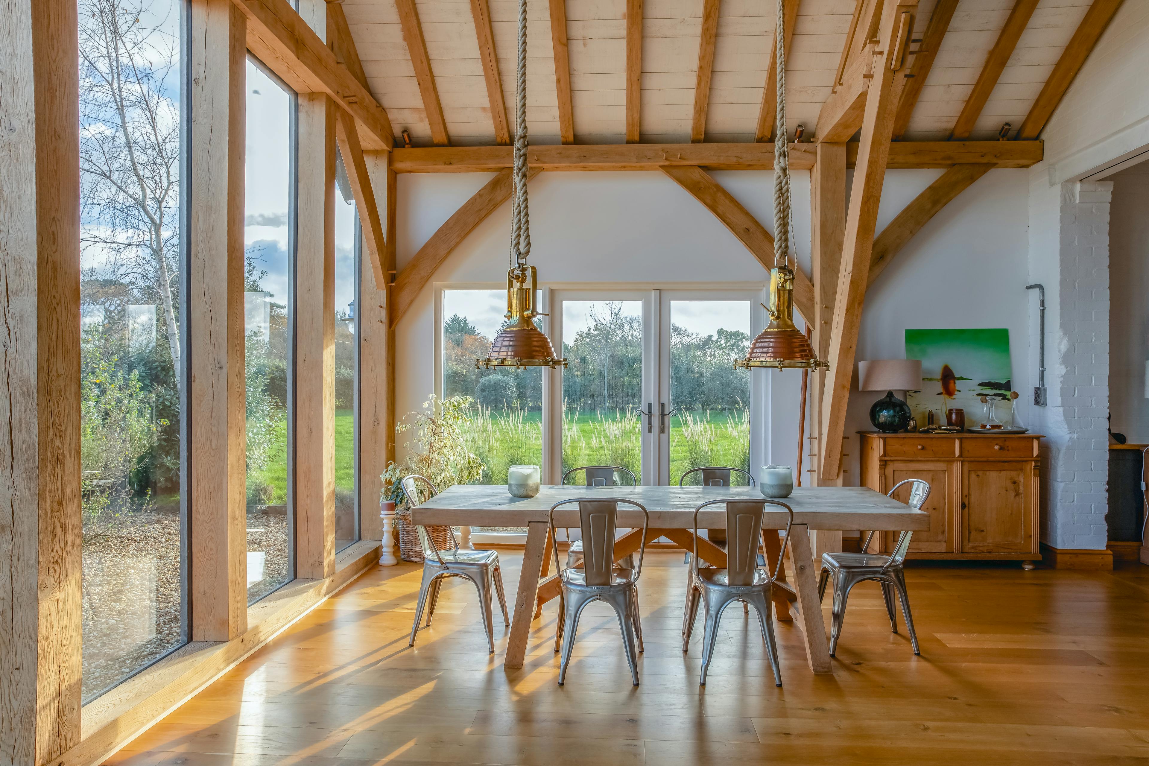 Sun beams shine through the windows on a wooden table and chairs and sideboard in an oak framed extension with a glazed gable