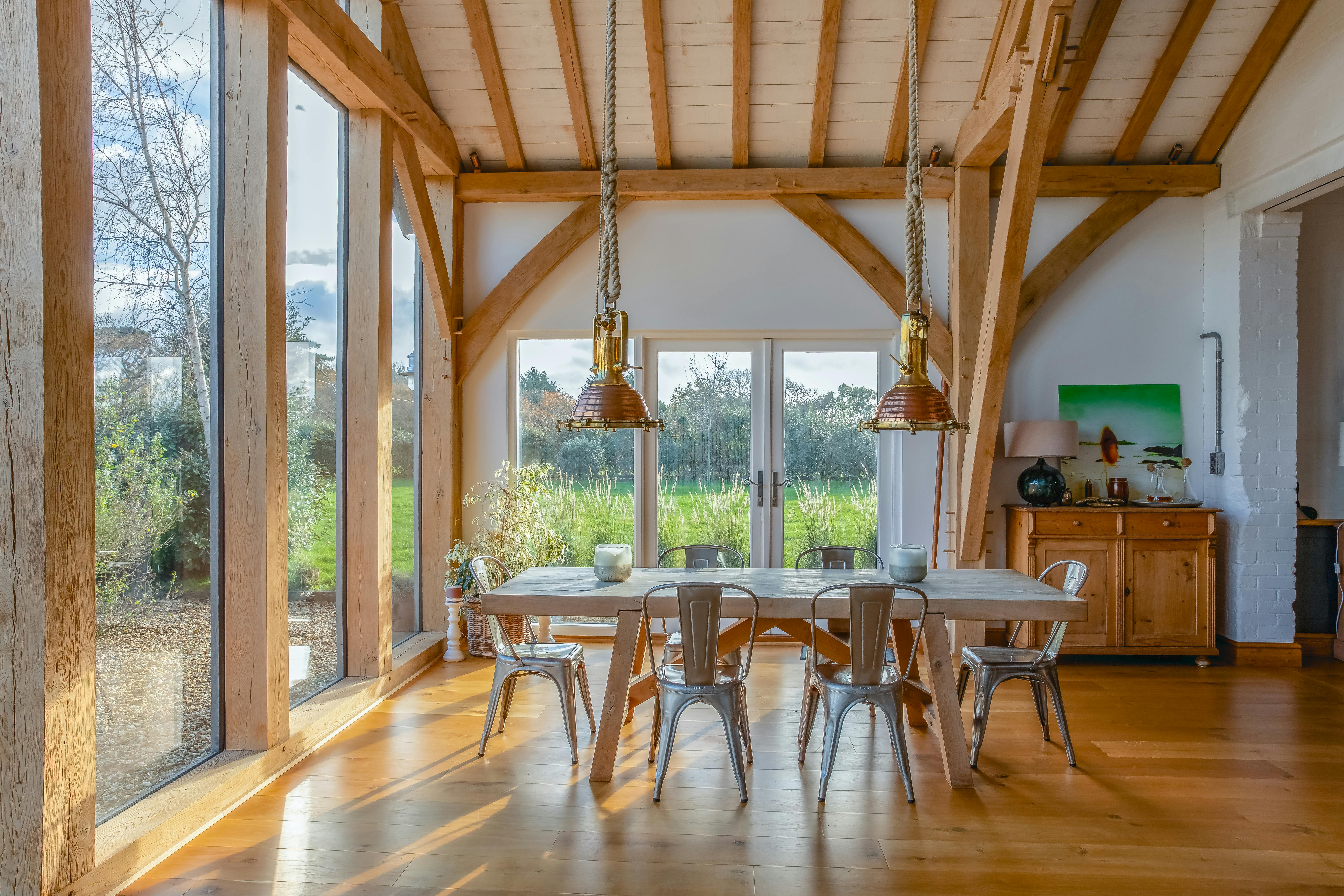 Sun beams shine through the windows on a wooden table and chairs and sideboard in an oak framed extension with a glazed gable